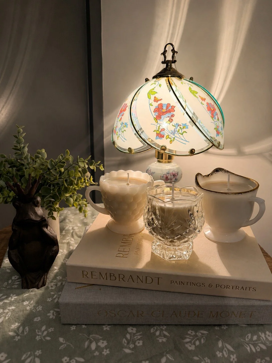 A decorative table setting with a floral lamp, three candles in unique holders, a potted plant, and two art books on a lace tablecloth.