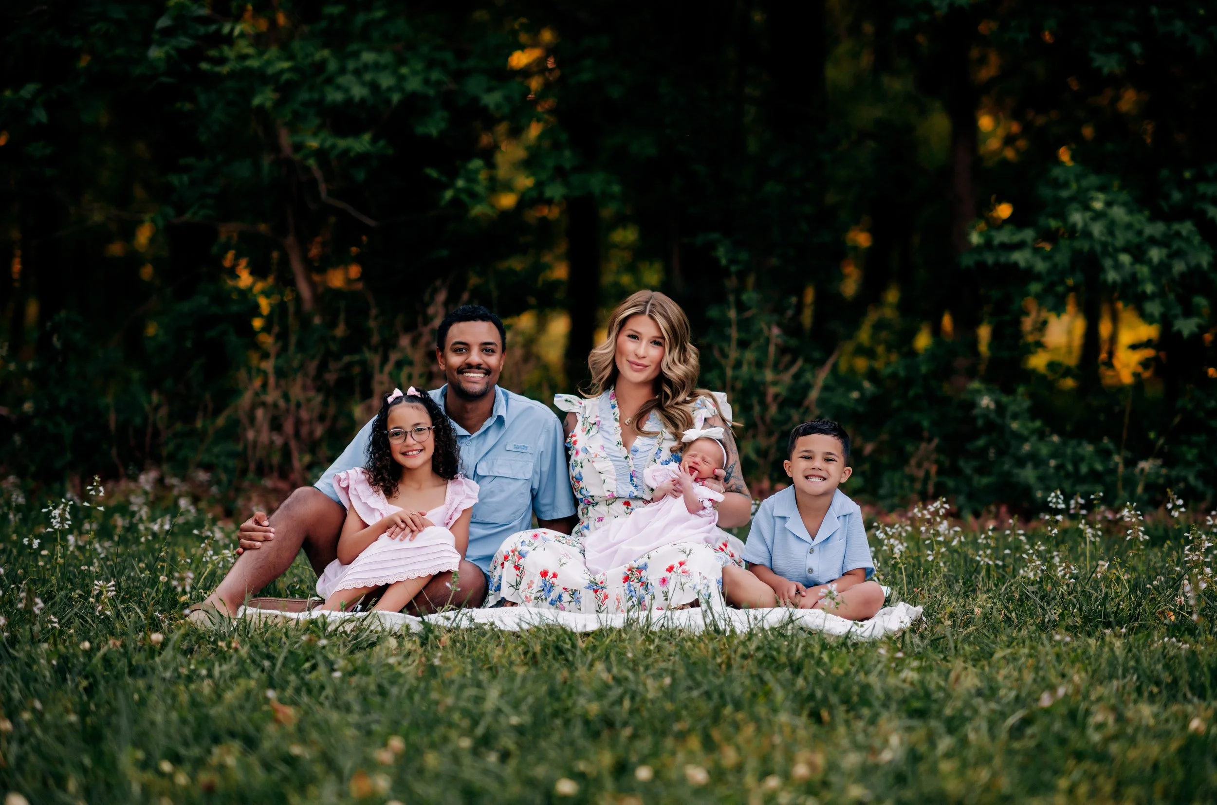 A diverse family of six sitting on grass in a park, smiling and posing for the camera with trees in the background.