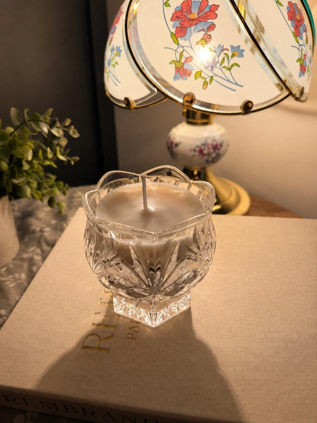 A decorative crystal glass candle holder with a white candle on a beige box, a floral table lamp in the background, and a small green plant to the left.