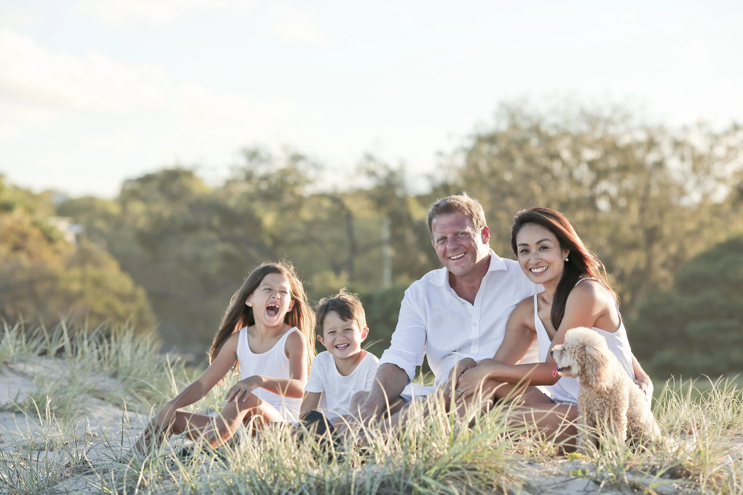 Un groupe familial souriant de cinq personnes dont deux adultes et trois enfants, assis sur la plage avec de l'herbe et un chien Chow Chow blond, au coucher du soleil avec un arrière-plan de arbres et ciel clair.