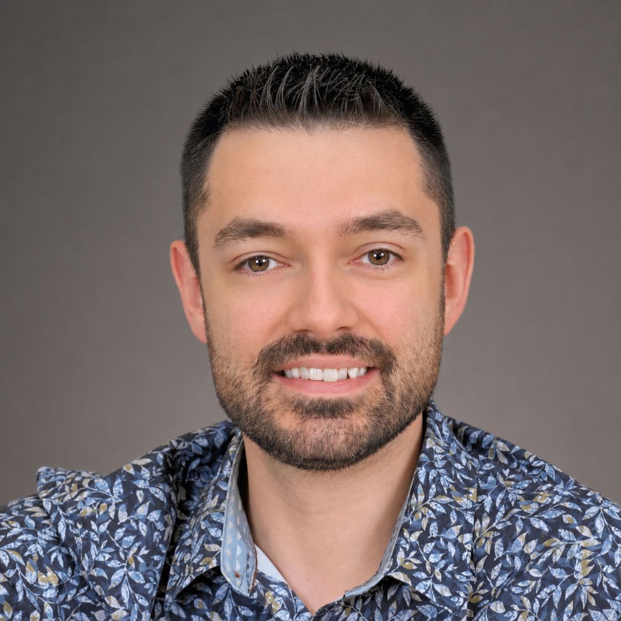 Un portrait d'un homme souriant avec une barbe, portant une chemise à motif floral sur fond gris.