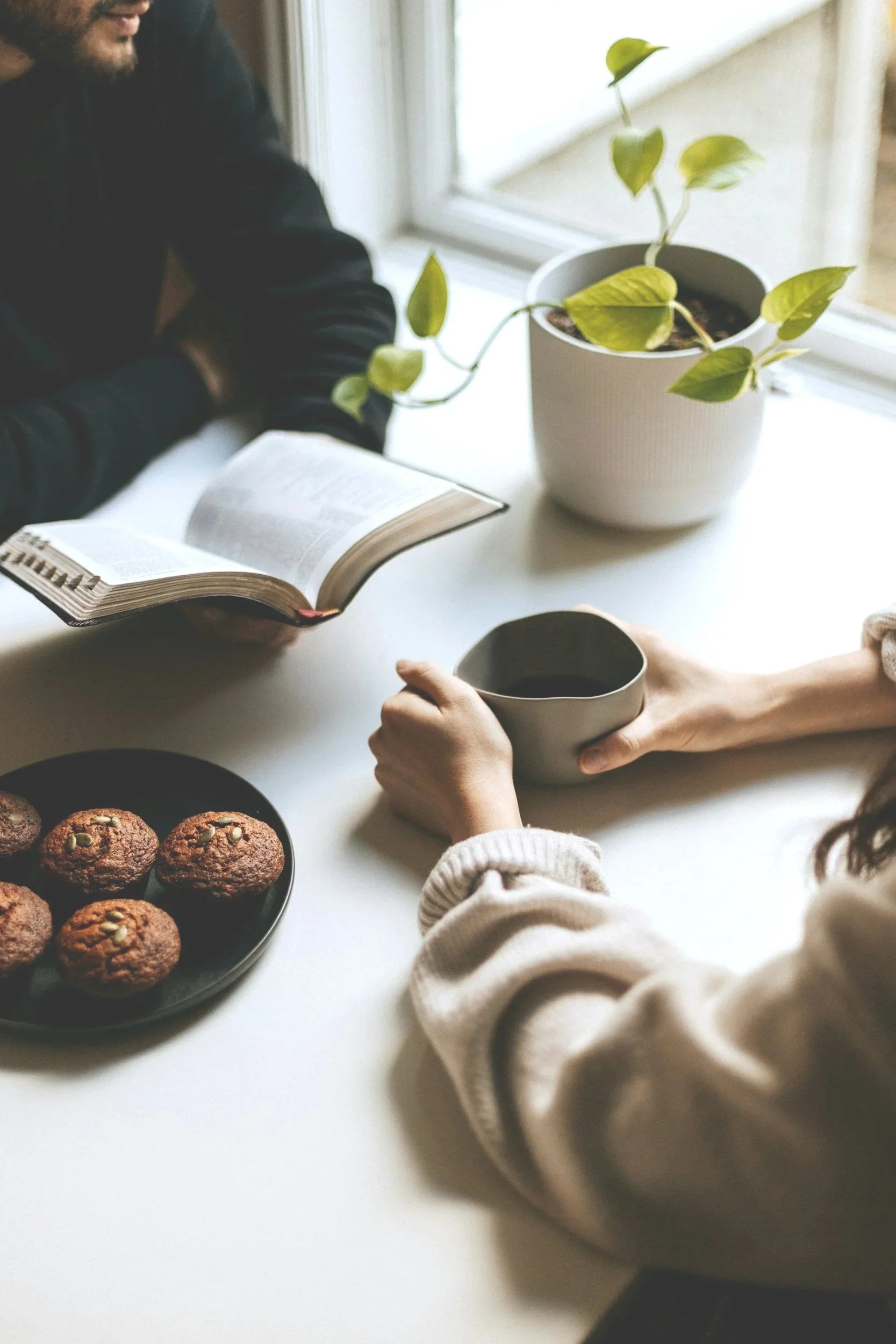 Deux personnes sont assises à une table avec un livre ouvert, une tasse, et un panier de muffins ou cookies. Une plante verte en pot est placée en arrière-plan près de la fenêtre