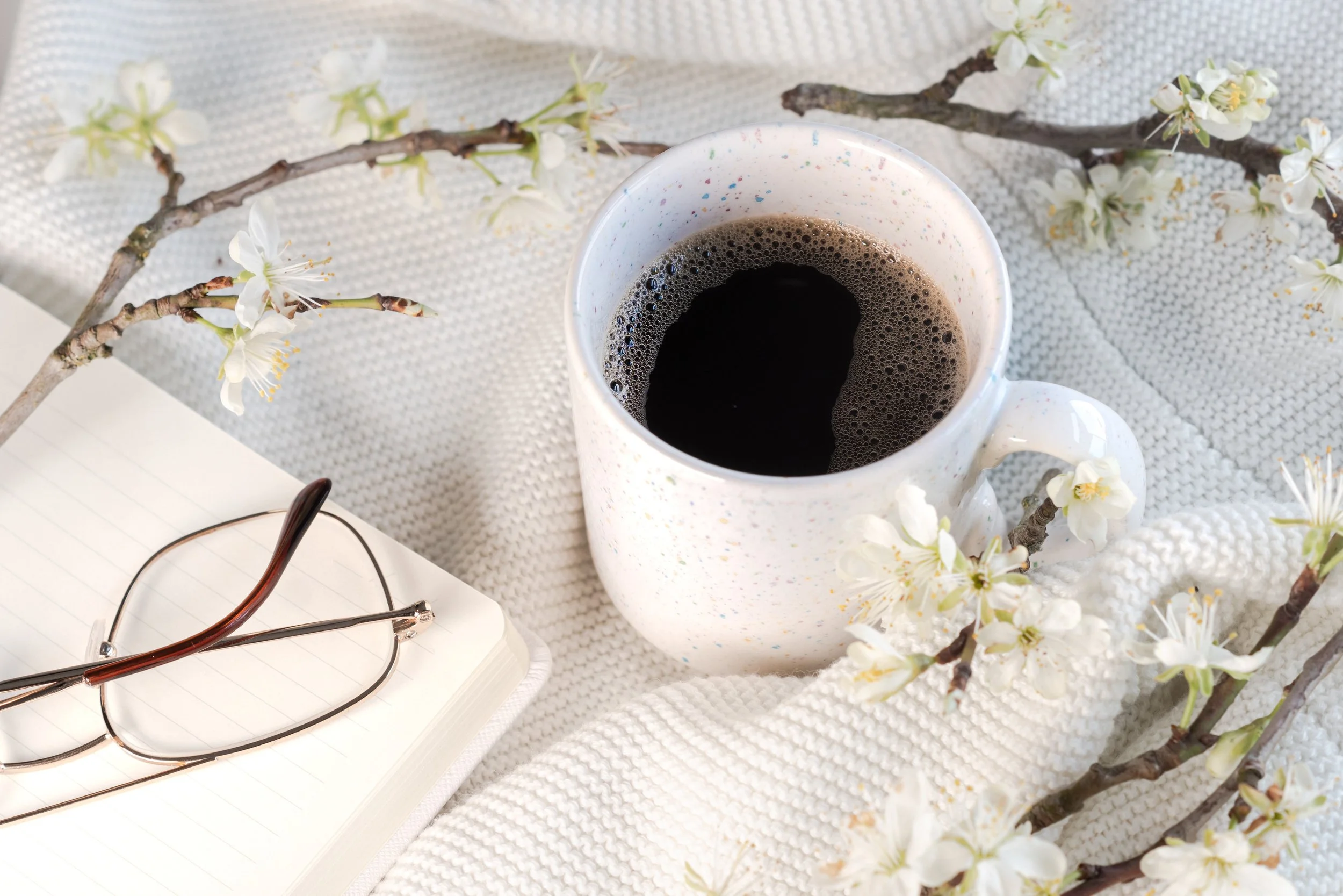 Une tasse de café noir, des branches de fleurs blanches, un carnet avec des lunettes dessus, et un espace pour écrire. Background avec une texture en tissu blanc.