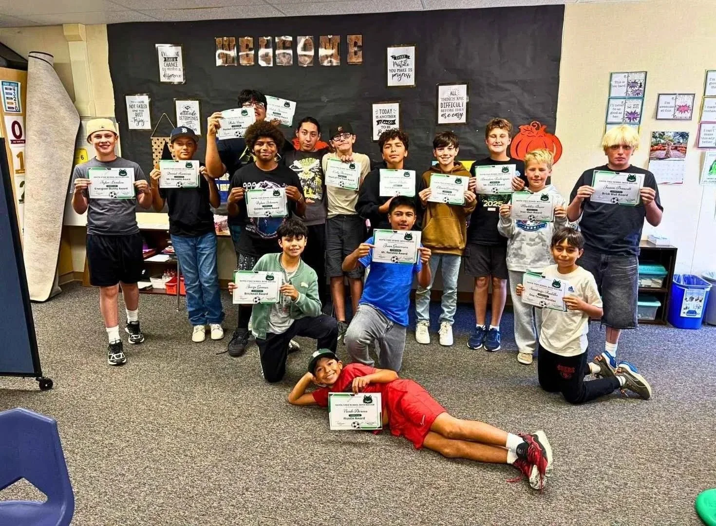 A group of children in a classroom holding certificates of achievement, with some kneeling or lying on the floor. The background has a blackboard with posters and a pumpkin decoration, indicating a classroom setting and a possible celebration.