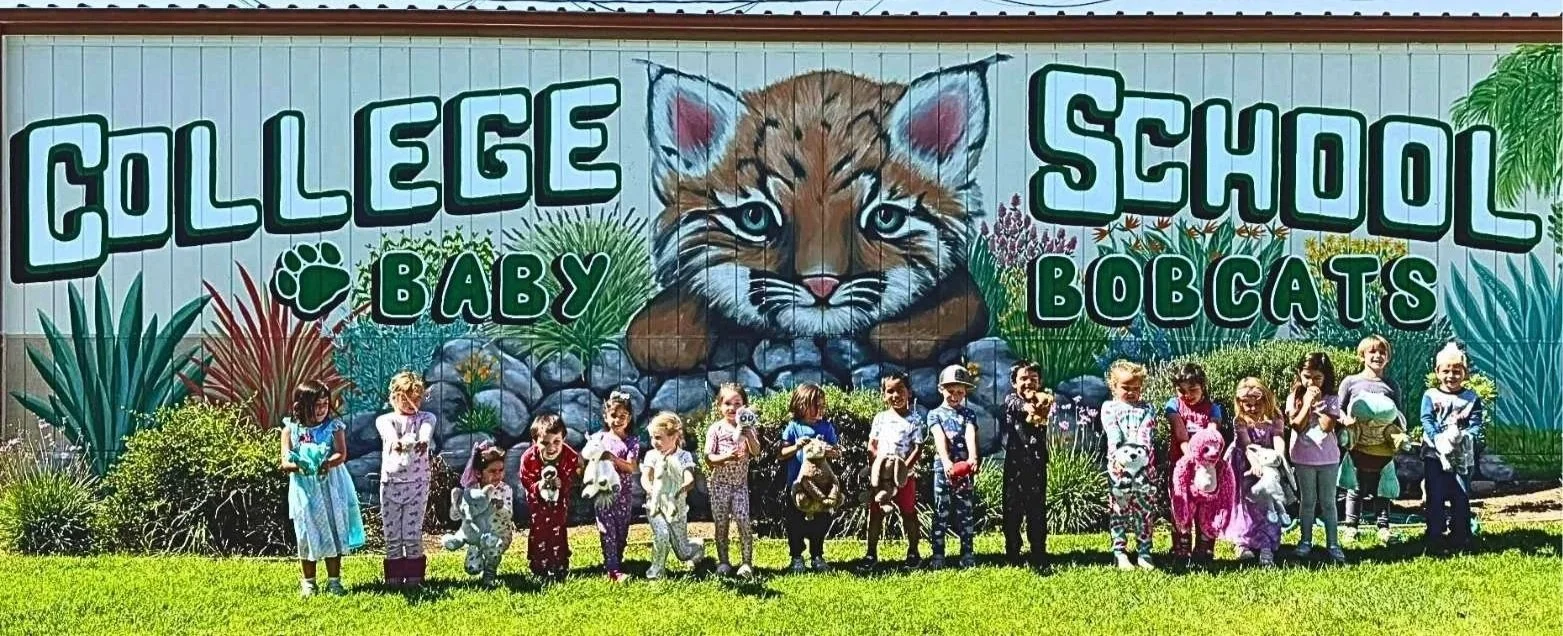 Group of children in TK and Kindergarten on the College Elementary School campus dressed in pajamas holding stuffed animals, standing in front of a mural that reads 'College Baby Bobcats' with a large painted bobcat face and surrounding plants.