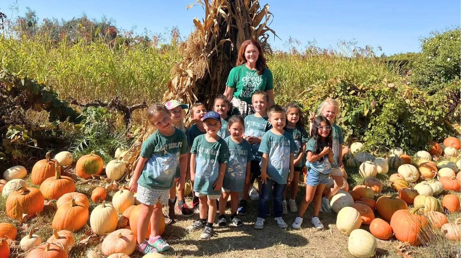 Group of children and their teacher at a pumpkin patch on a sunny day, surrounded by pumpkins and corn stalks.