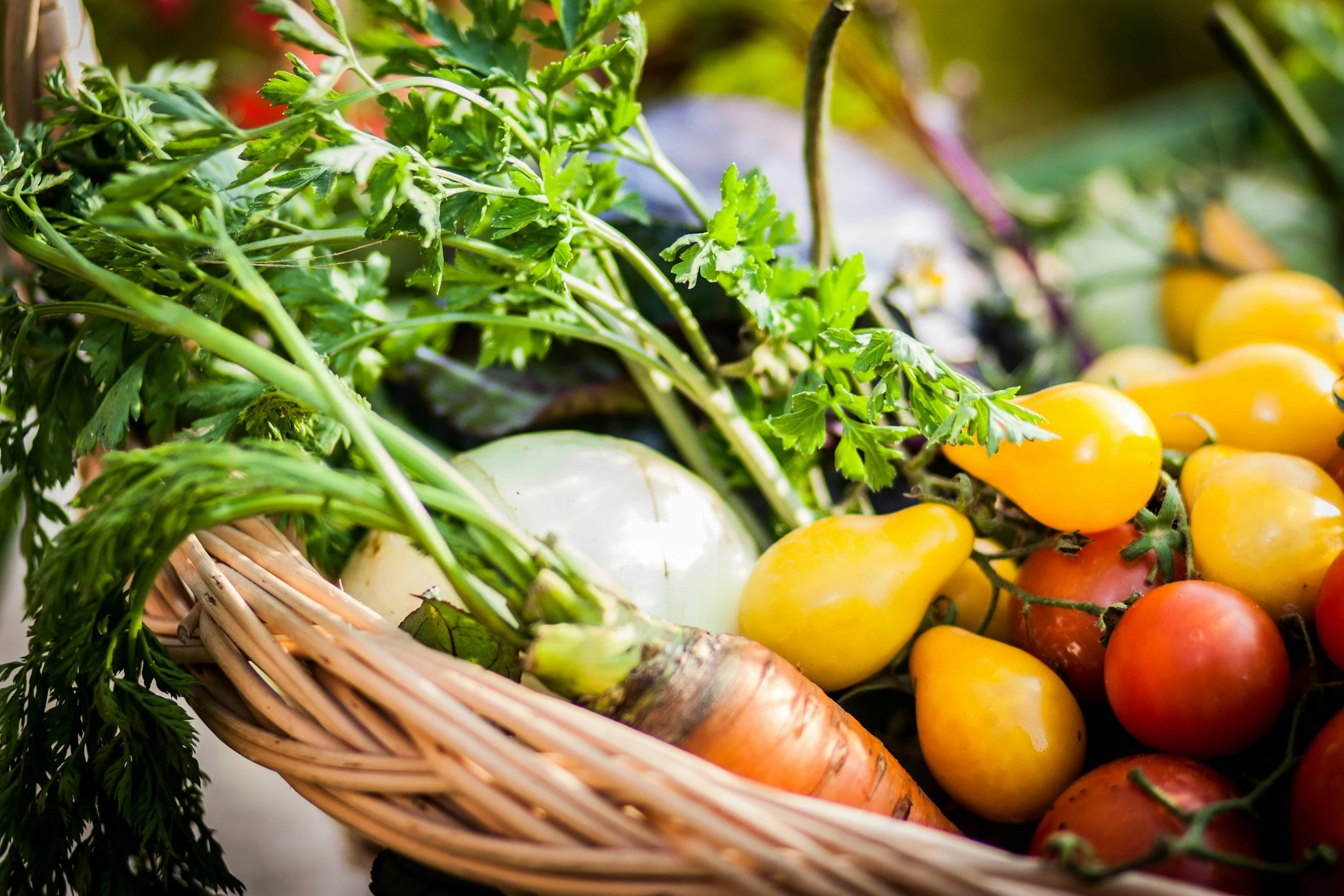 A close-up of fresh vegetables in a wicker basket, including green leafy herbs, white onion, orange carrot, and multicolored cherry tomatoes.