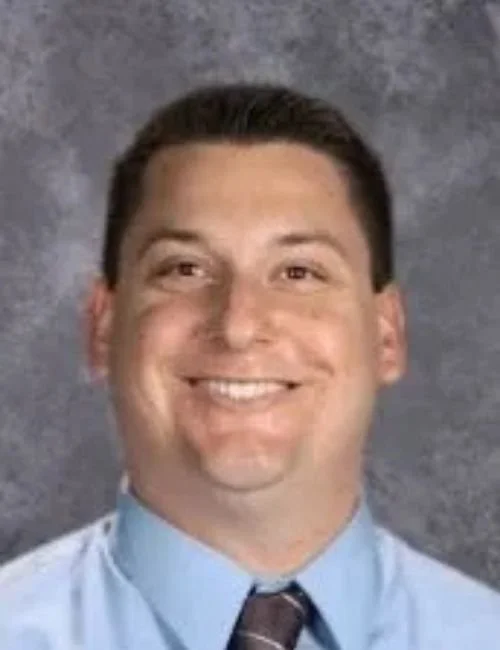 Principal Anthony Bruenner smiling, wearing a light blue dress shirt and a dark tie, against a gray background.