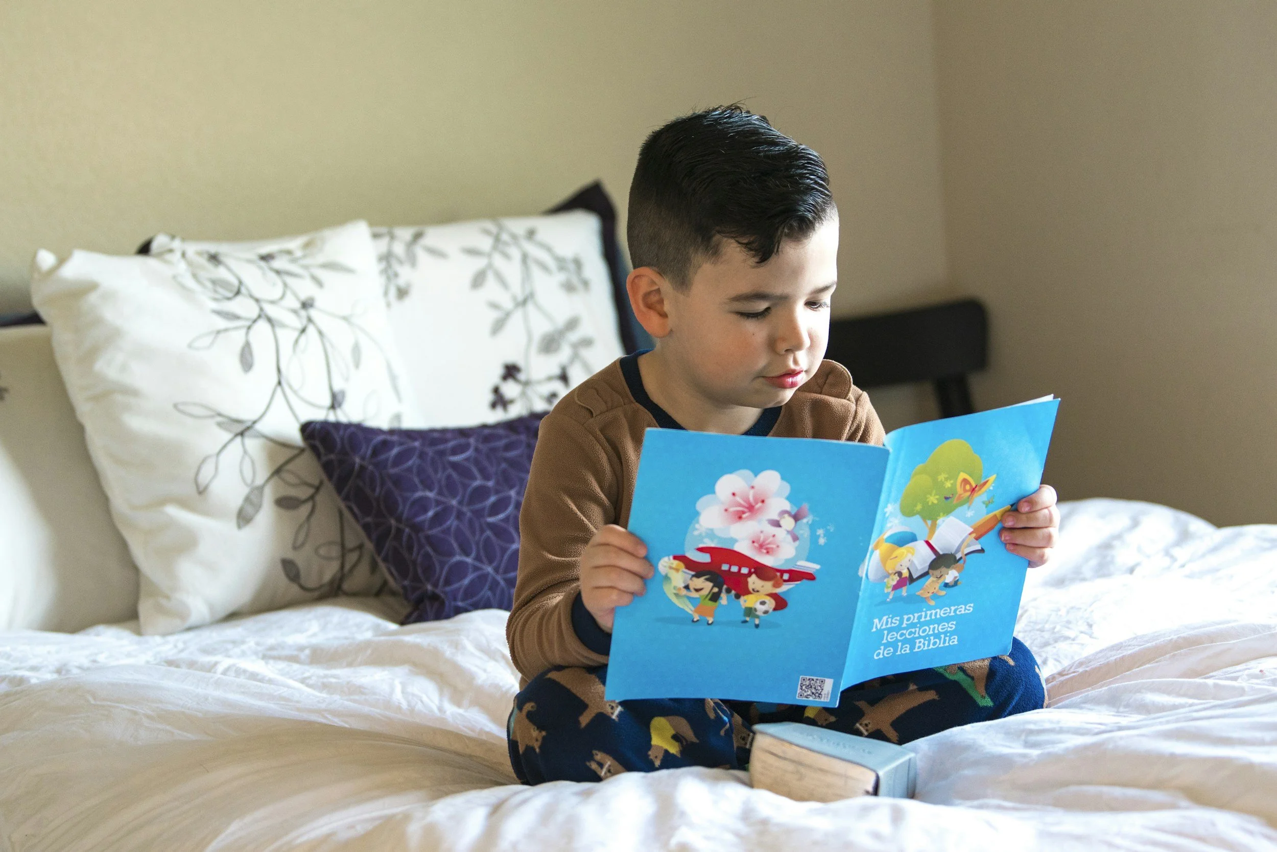 A young boy sitting on a bed, reading a colorful children's book titled 'Mis primeras lecciones de la Biblia,' with pillows behind him and beige walls in the background.