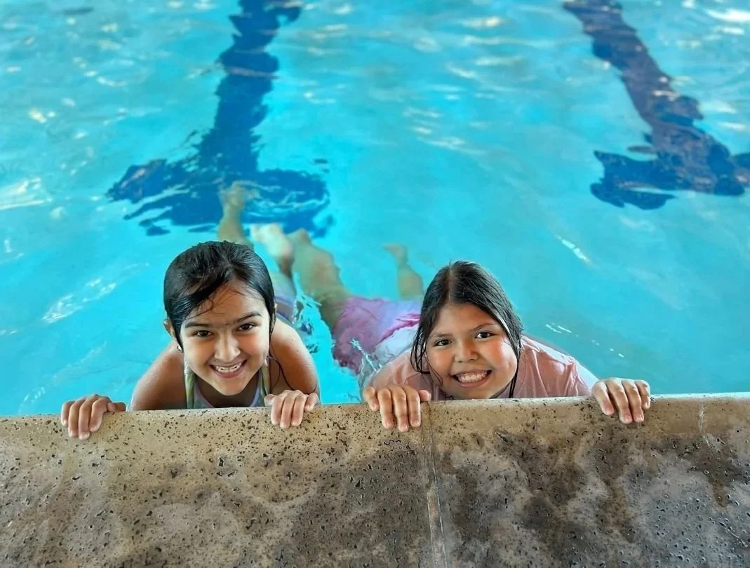Two young girls are smiling and holding onto the edge of a swimming pool while floating in the water.