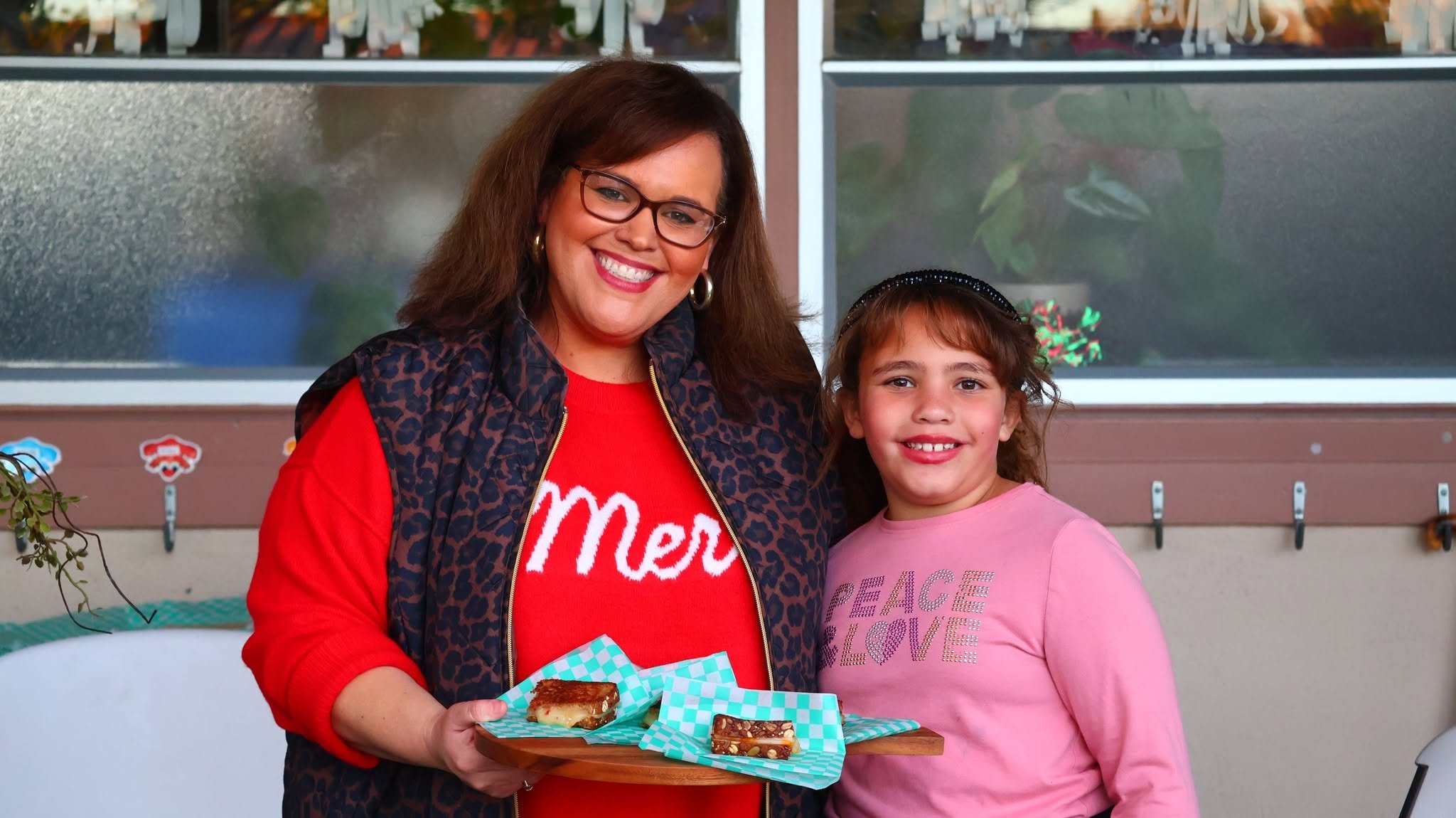 A woman and a young girl standing close together outdoors, smiling for the camera. The woman is holding a plate with dessert, possibly brownies or cakes, and they are both dressed casually. The woman is wearing glasses, earrings, and a red shirt with a leopard print jacket. The girl has curly hair, a black headband, and a pink shirt with printed words.