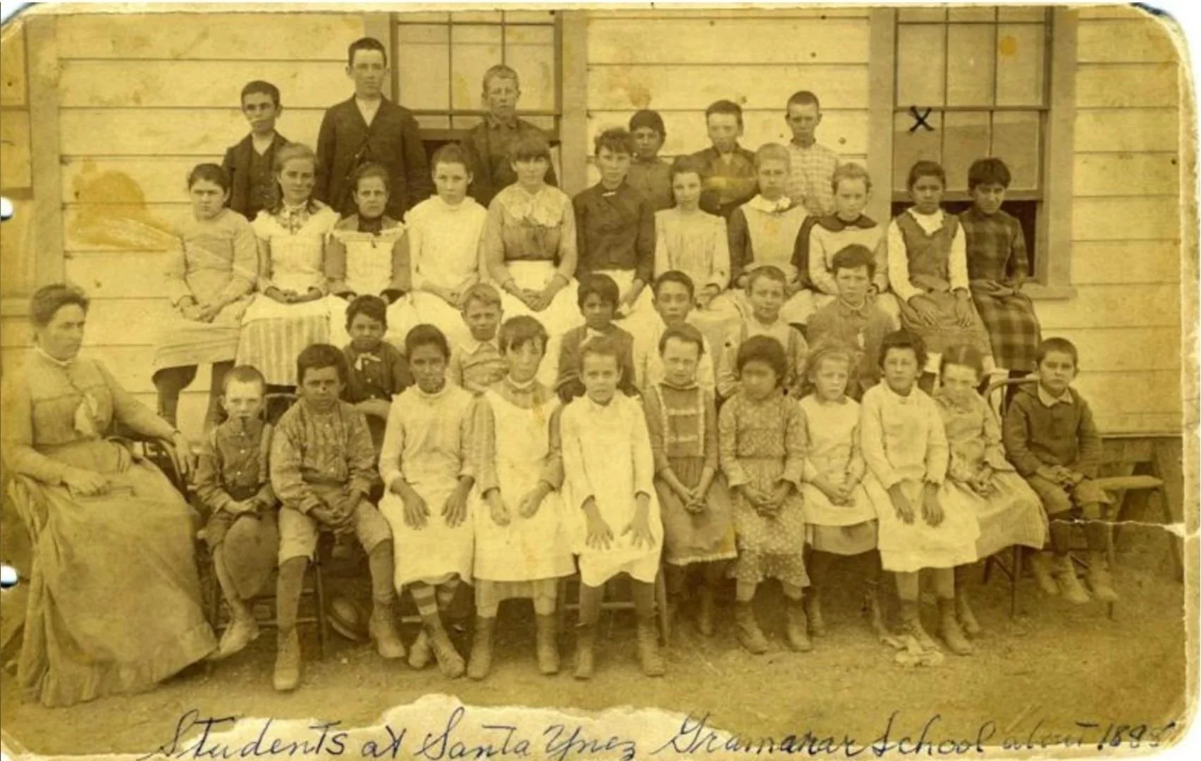 Group of young students and teacher sitting and standing in front of a wooden school building, posed for a class photo. The photo appears old, sepia-toned, with handwritten text at the bottom reading "Students at Santa Ynez Grammar School."