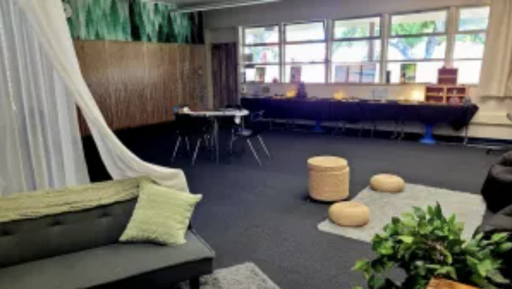 A spacious classroom with large windows, a black couch with a green pillow, a white hanging chair, a patterned rug with three beige round stools, a plant, and a long table along with some chairs and educational materials.