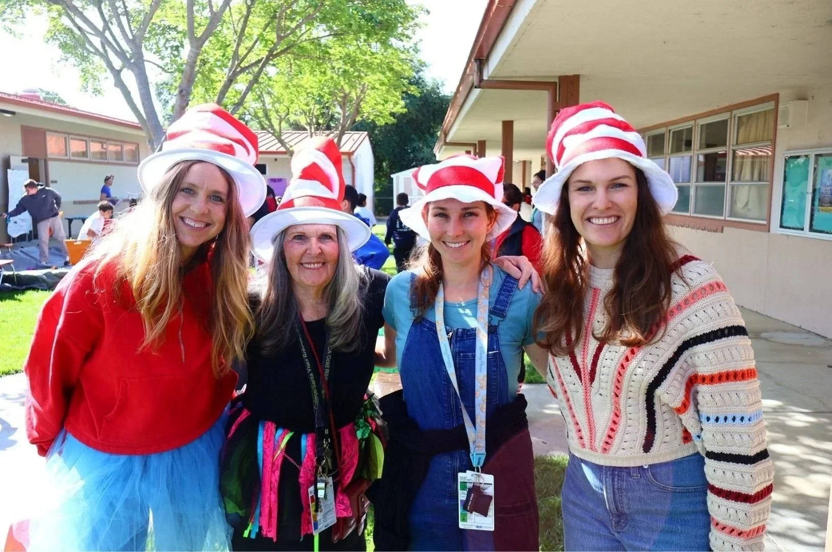 Four women wearing red and white striped hats inspired by the Dr. Seuss character, standing outdoors with school buildings and trees in the background.