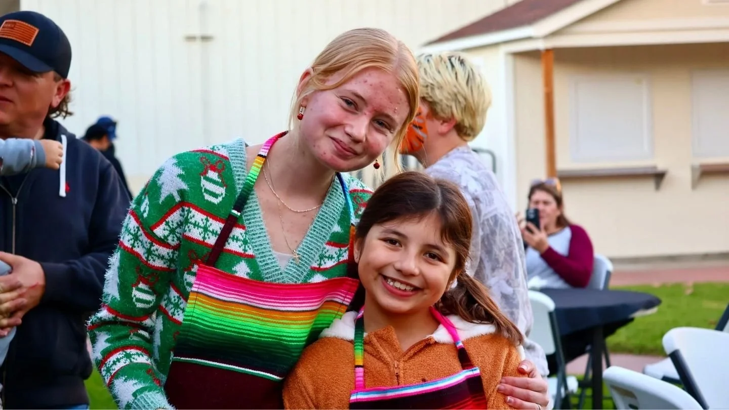 Two women, one with blonde hair and the other with brown hair, smiling and wearing festive Christmas sweaters, in an outdoor gathering. The blonde woman is wearing a colorful apron, and the girl has a colorful strap. In the background, other people are seen, including a woman taking a photo.