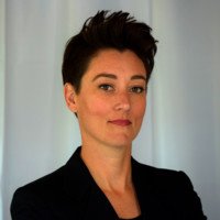 Photo of the Board President, Calisse Courtney, wearing a black blazer, standing in front of a light background.
