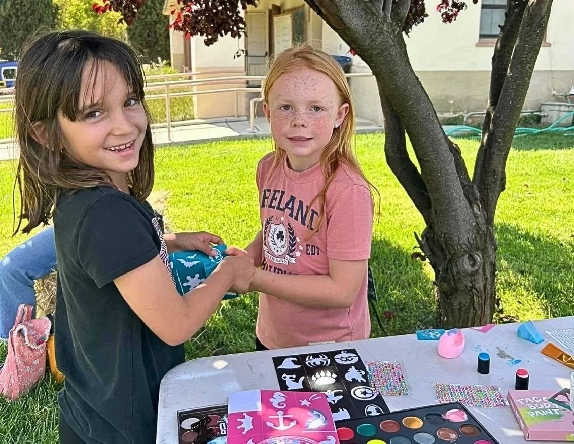 Two young girls are outdoors under a tree, exchanging a wrapped gift. The table in front of them holds face paint kits, stickers, and craft supplies. The girl on the left has dark brown hair and is wearing a black T-shirt, and the girl on the right has red hair, freckles, and is wearing a pink T-shirt.