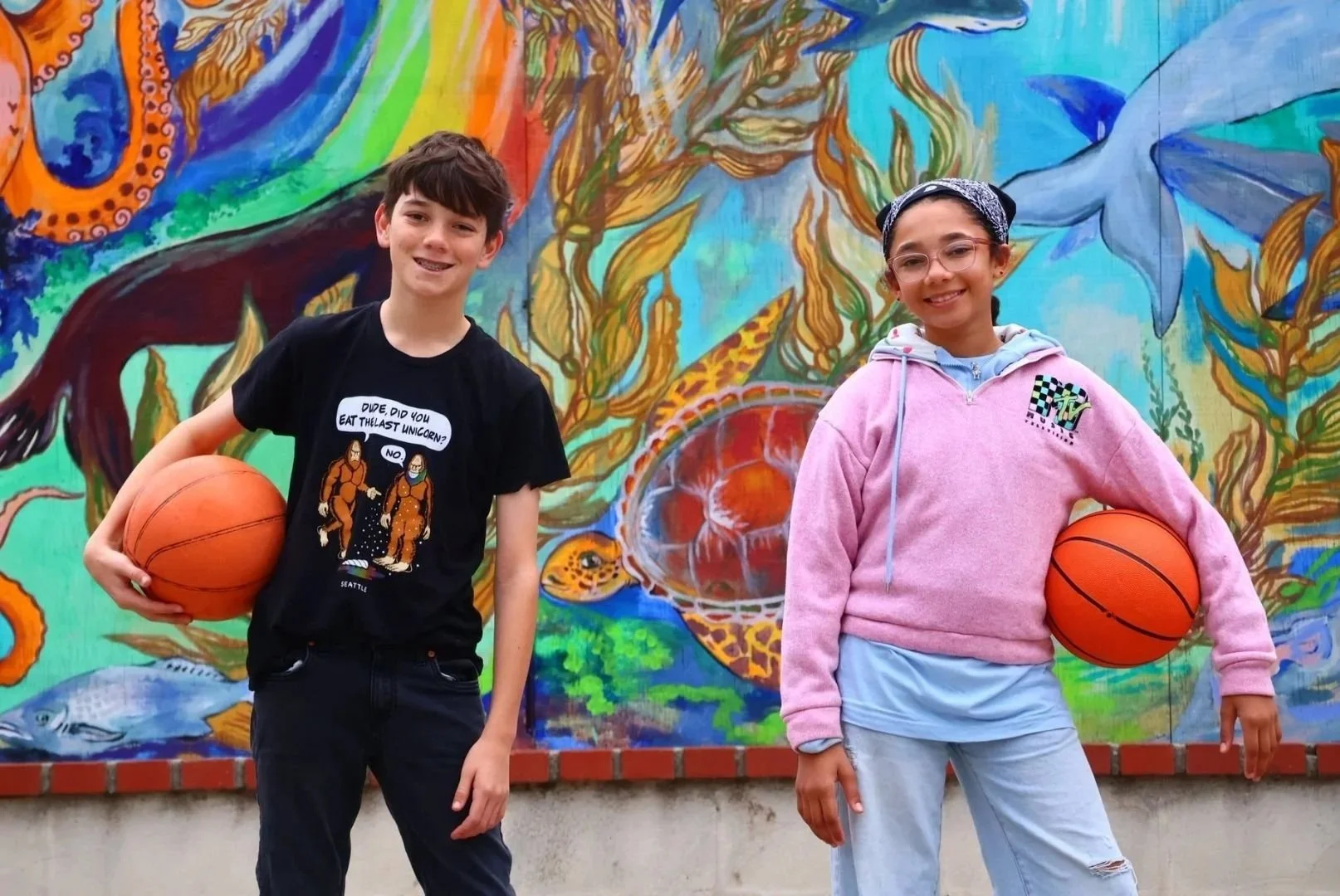 Two children standing in front of a colorful mural holding basketballs. The boy on the left is wearing a black T-shirt with cartoon characters and text, and the girl on the right is wearing a pink hoodie and glasses.
