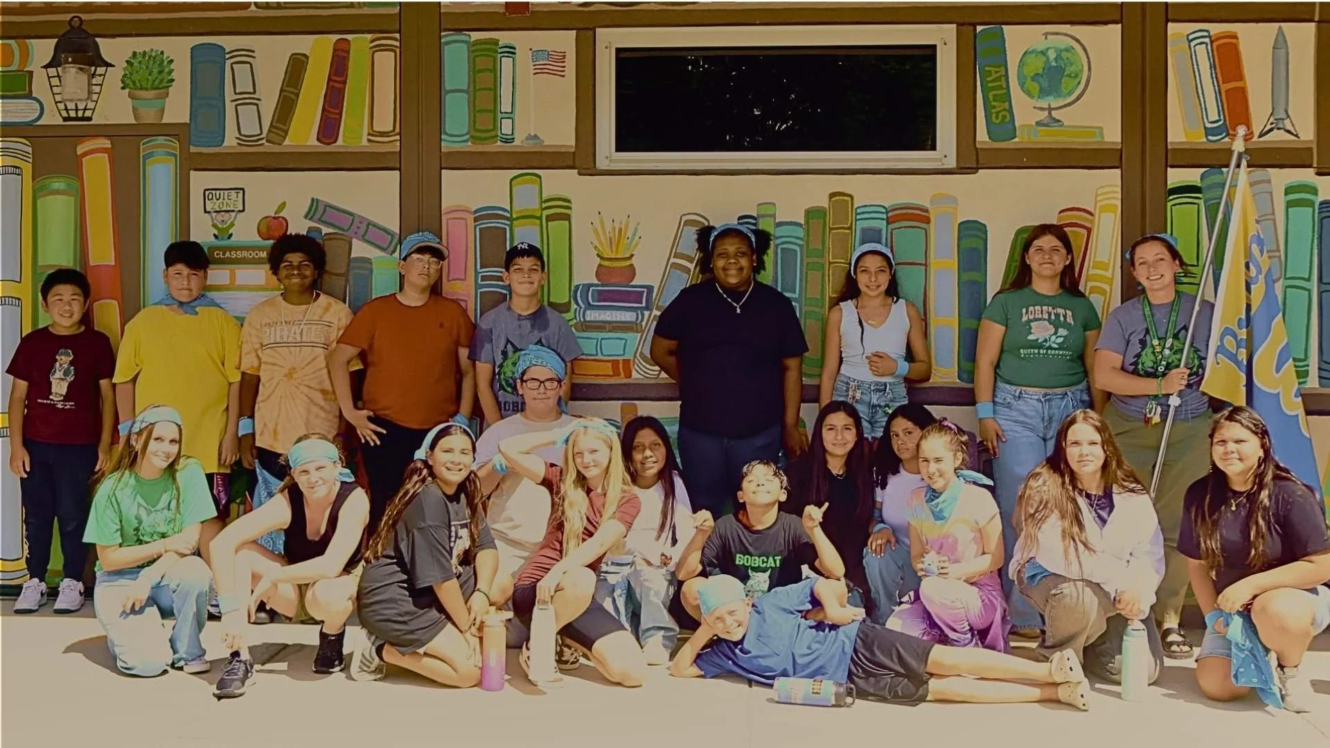 A group of children and adults posing in front of a colorful mural of books and a globe, some children sitting and some standing, with two adults holding flags.