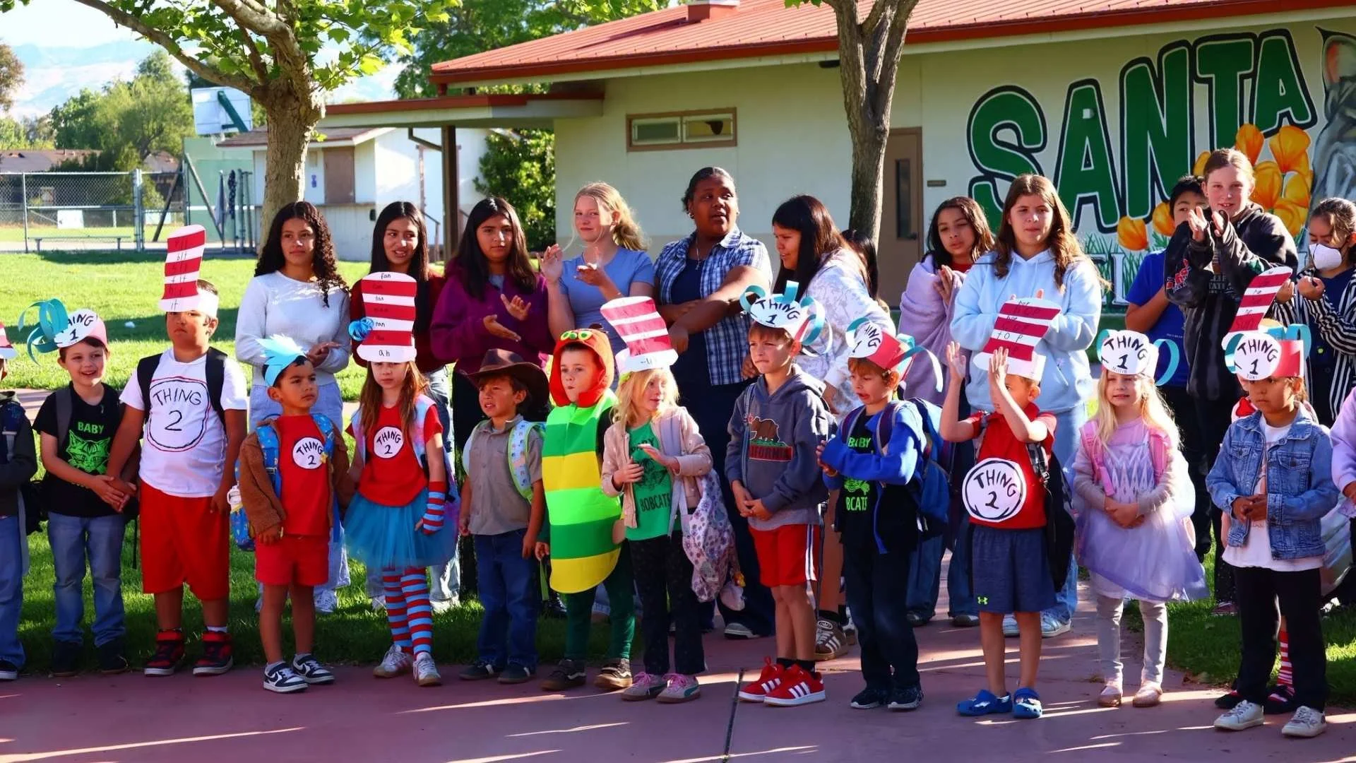 A group of children and adults gathered outside on a grassy area with trees, participating in a school or community event. Some children wear hats labeled 'Thing 1' and 'Thing 2,' and costumes inspired by Dr. Seuss. The background features a building with a colorful mural reading 'Santa' and a basketball hoop. The children are standing on a paved surface, with the sun shining.