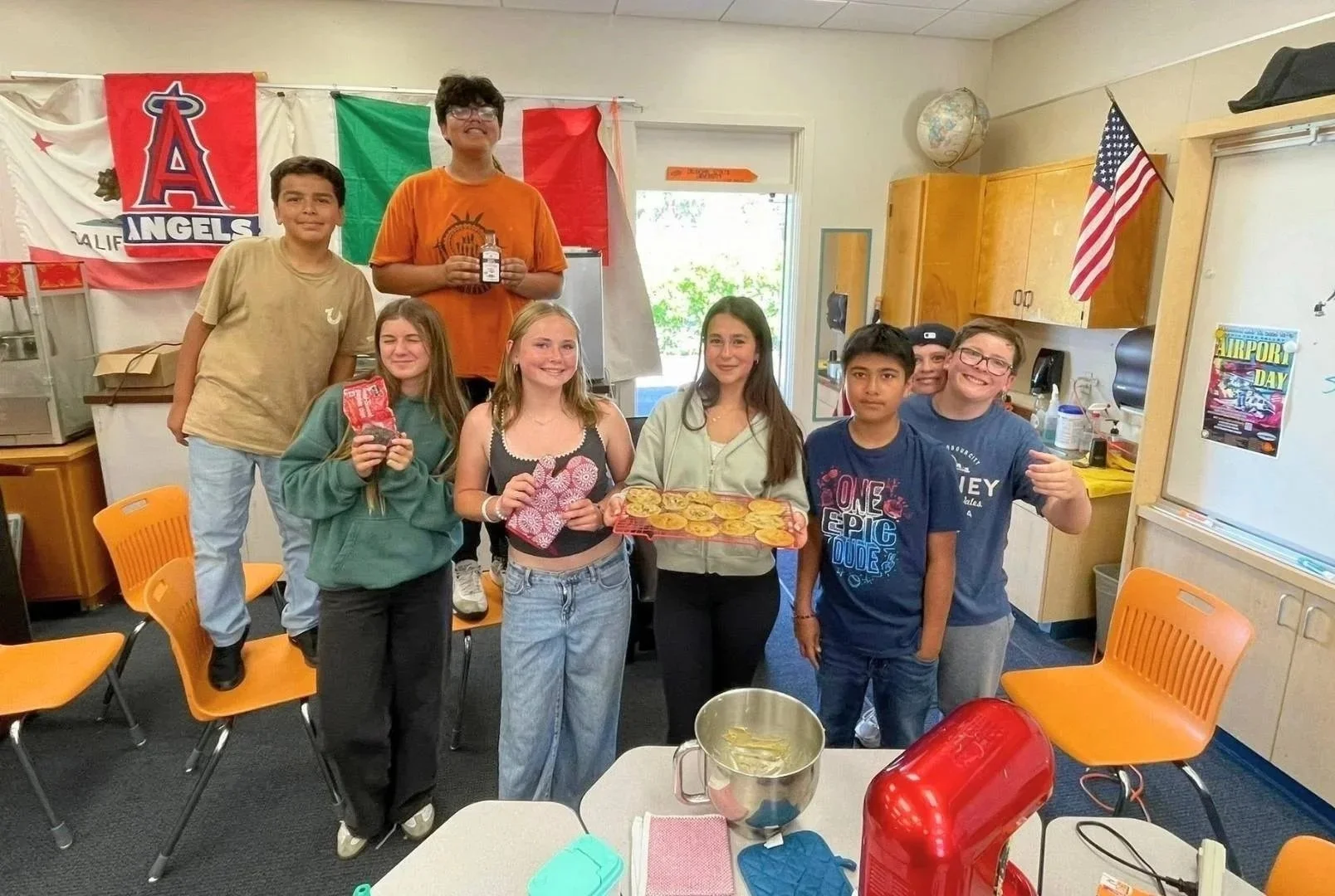 A group of children in a classroom celebrating airport day, holding cookies and snacks, with flags and banners in the background.