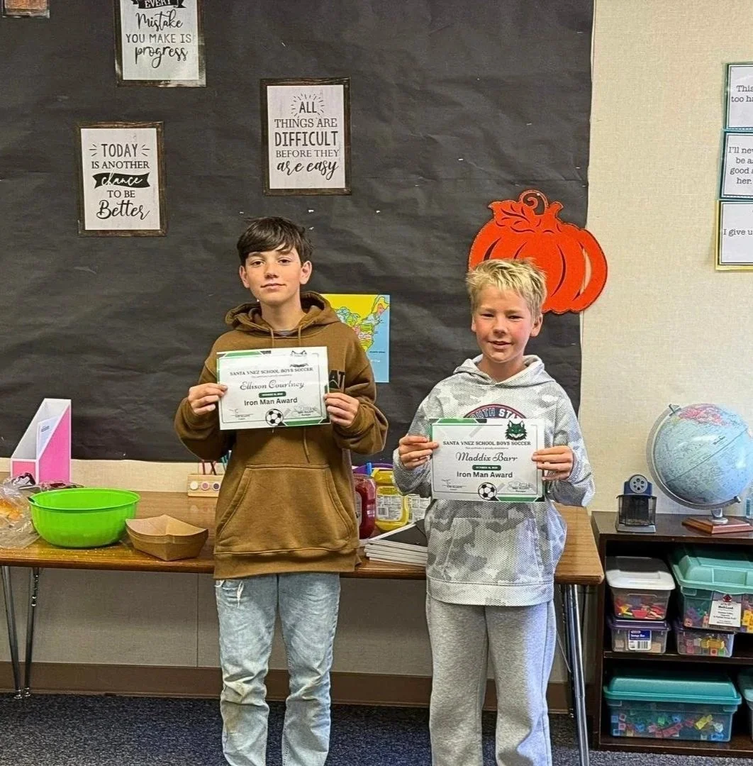 Two boys standing in a classroom holding certificates for the Iron Man Award. The boy on the left has dark hair and is wearing a brown hoodie, while the boy on the right has blonde hair and is wearing a gray camouflage hoodie. Behind them is a black bulletin board with motivational posters and an orange pumpkin decoration. There are supplies on the table behind them, including a green bowl and paper trays.