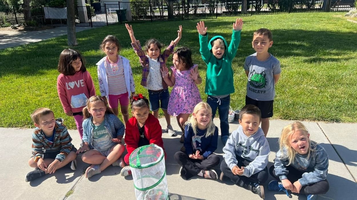 Group of children outdoors on a sunny day, some standing with hands raised, others sitting on the sidewalk, with a green grassy background and a fence.