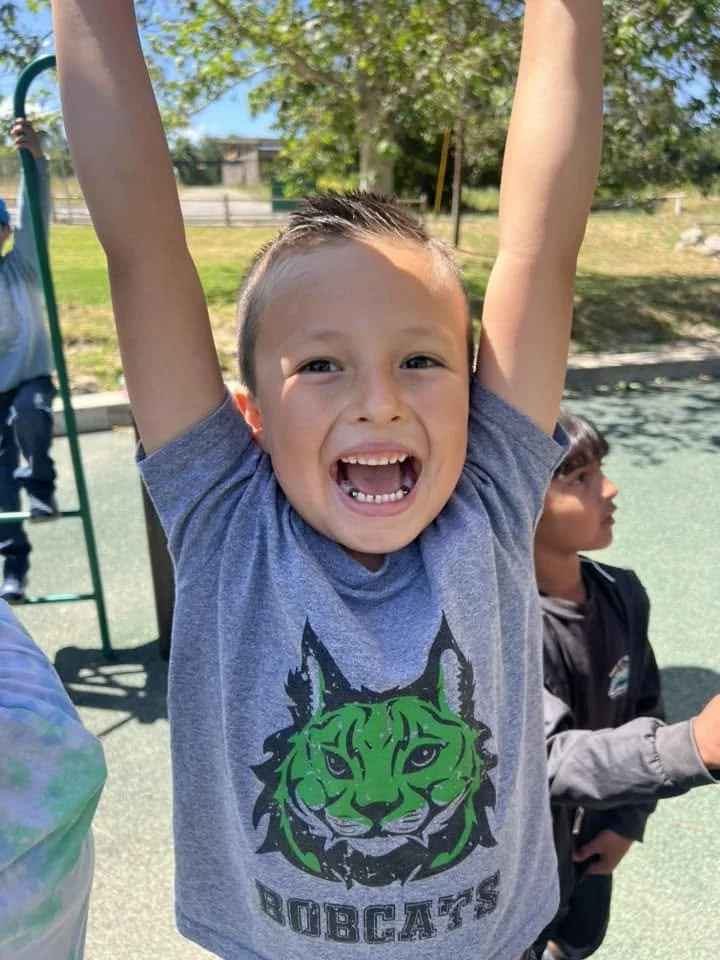 A young boy is smiling widely with arms raised, wearing a gray t-shirt with a green and black Bobcats logo, outdoors on a sunny day with two children in the background.