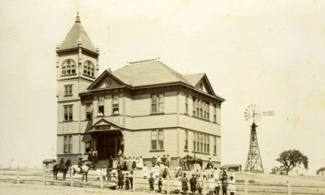 Old black and white photo of a large, historic College School District building with a tower, surrounded by people and horses, and a windmill in the background.