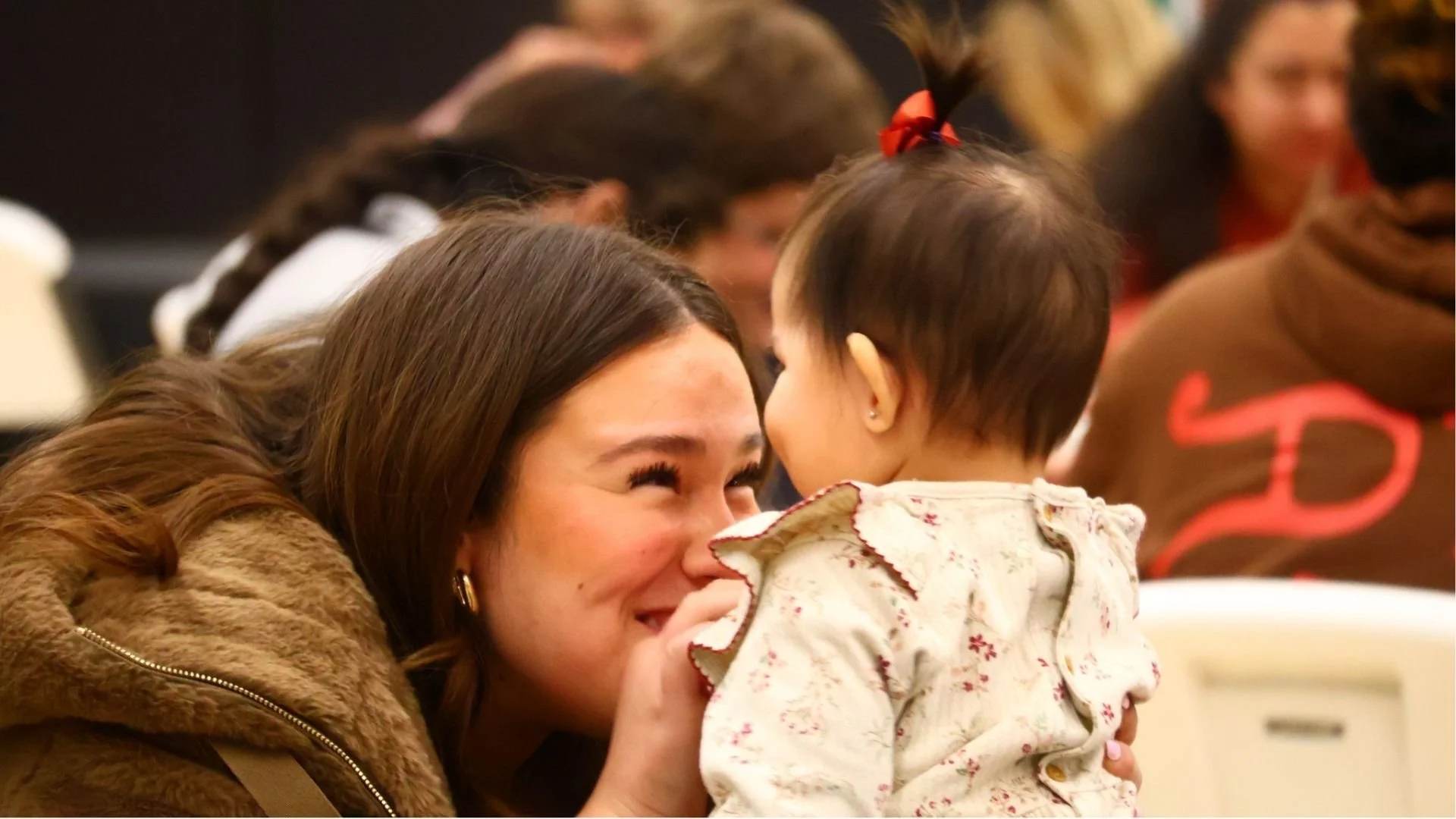 A woman and a young girl sharing a close moment, with the woman smiling and the girl facing her. The woman has long brown hair, and the girl has short dark hair with a red bow. People are blurred in the background.