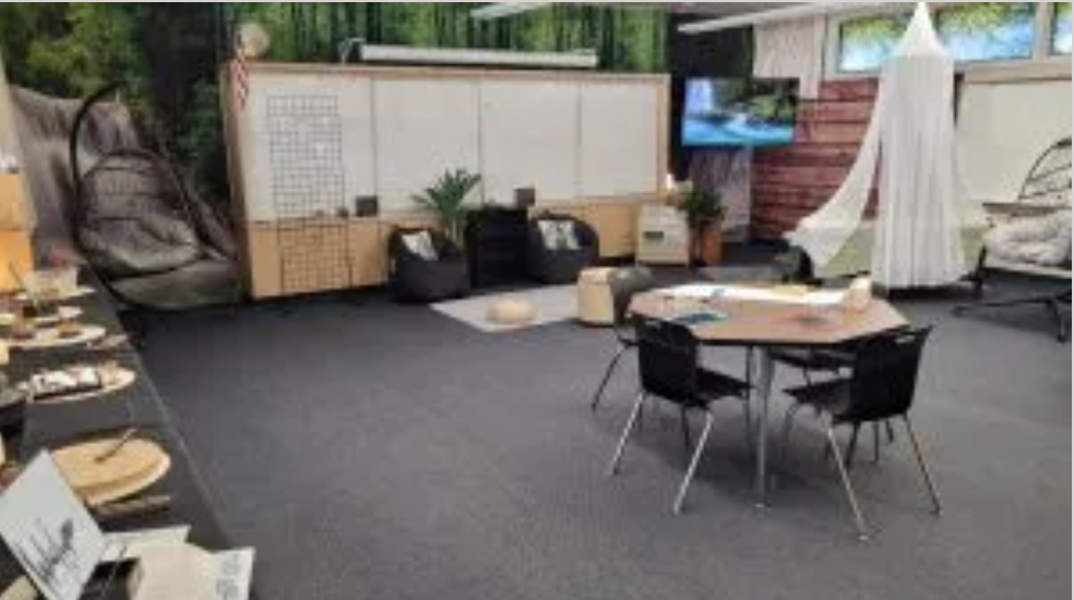 Indoor modern lounge with black armchairs, a round table with four black chairs, shelves with books, plants, and decor, and a large white fabric hanging. Back wall has windows with greenery outside.