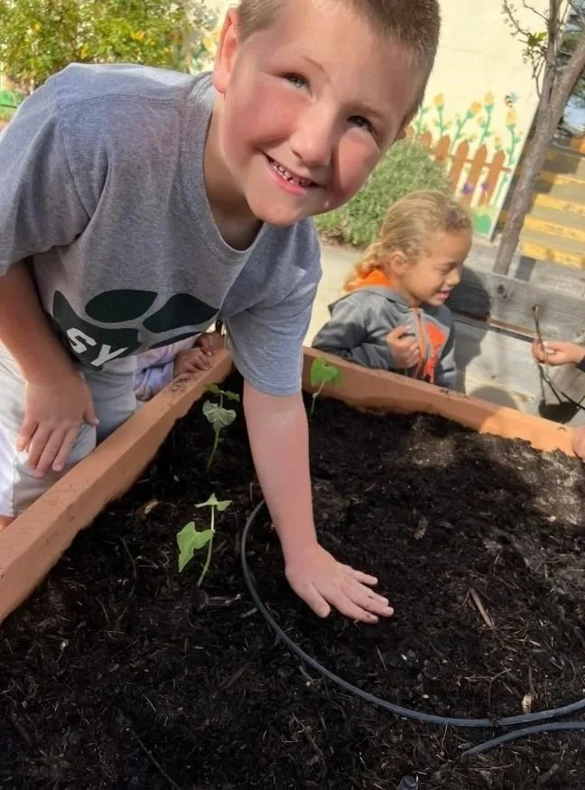 A young boy smiling while planting seedlings in a garden bed, with two other children in the background.