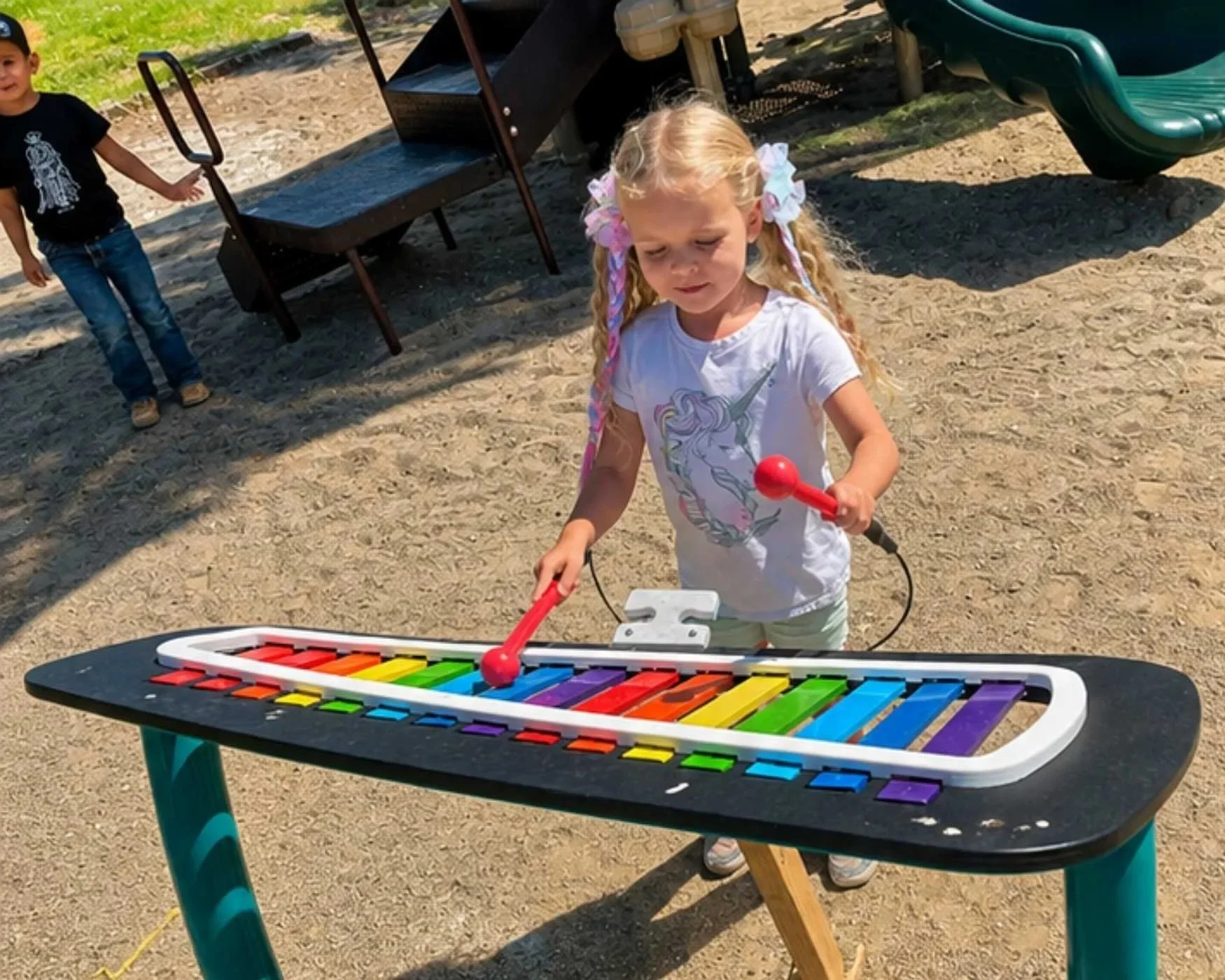 A young girl with blond hair and colorful bows, wearing a white unicorn t-shirt, is playing a rainbow-colored xylophone at an outdoor playground. A boy in a black t-shirt and jeans is standing in the background near playground equipment.