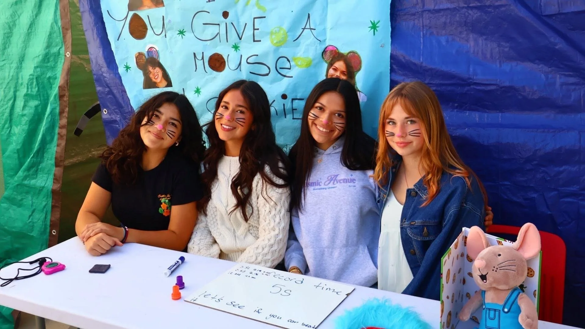 Group of four young women with face paint of cat whiskers and pink noses, sitting or standing behind a table at a lively outdoor event, with a colorful handmade sign that reads 'You Give A Mouse' in the background.