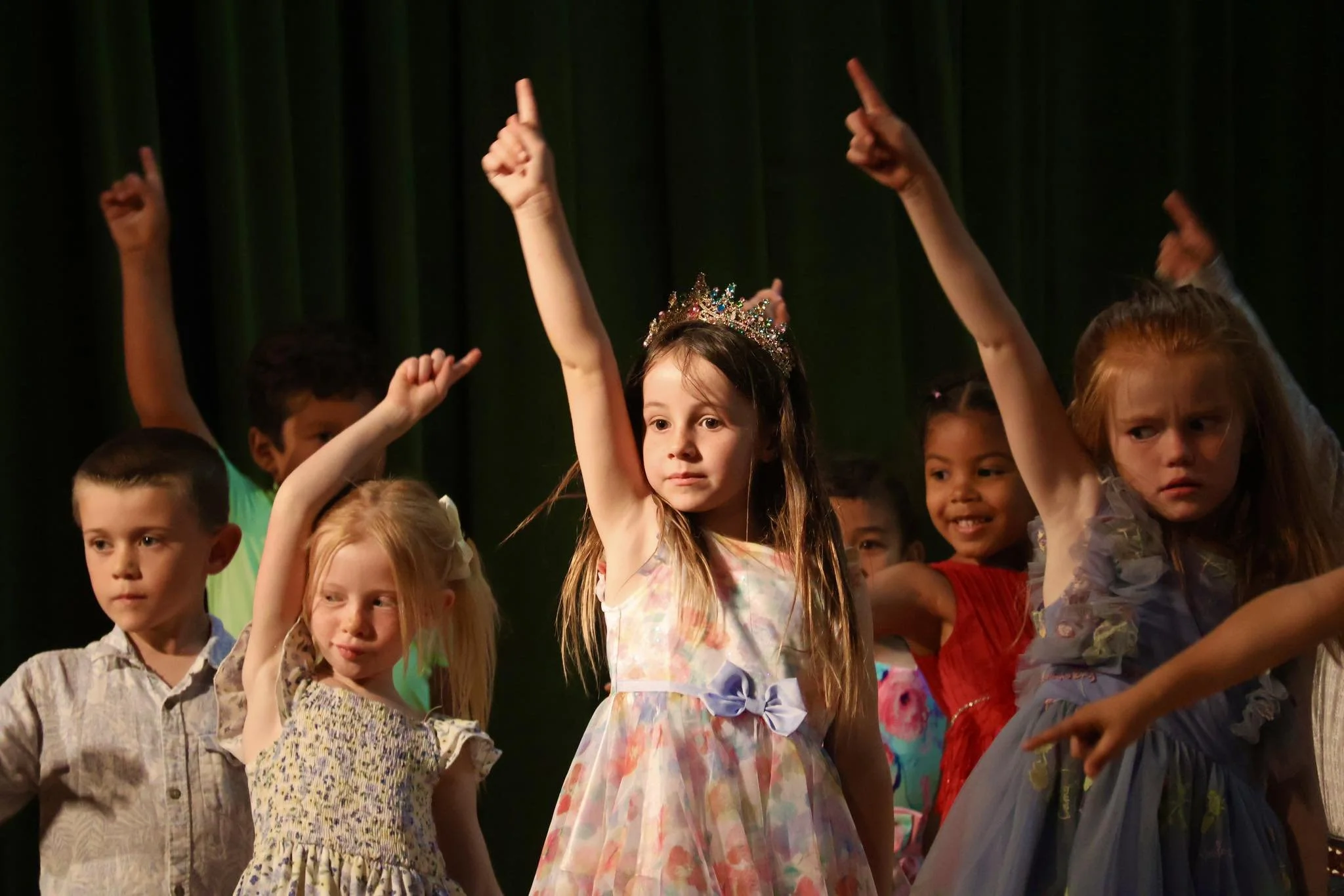 Young girl wearing a colorful dress and a crown performs on stage with other children during a school performance.
