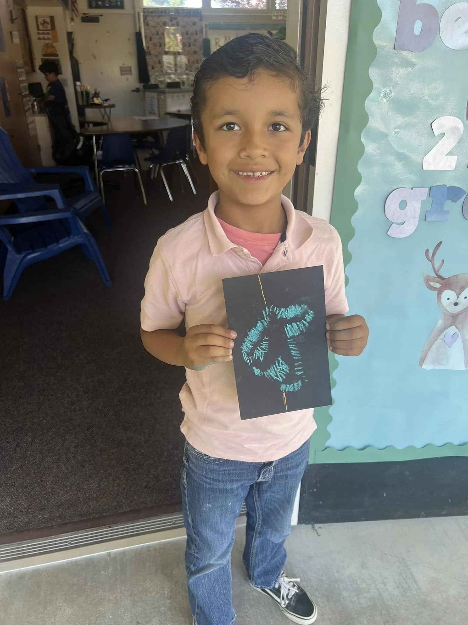 A young boy with dark hair and a big smile holding a black paper with a colorful chalk drawing of a flower. He is standing in front of a classroom door with a light blue border and holiday decorations. The classroom interior with desks and another child is visible in the background.