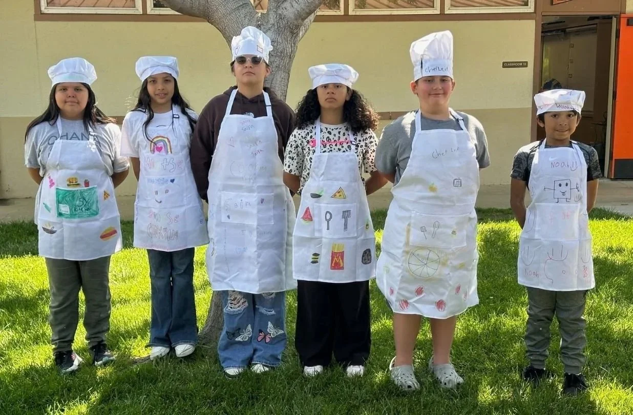 Six children standing outdoors in front of a tree, wearing homemade aprons and chef hats with drawings and names on them, with a school building in the background.