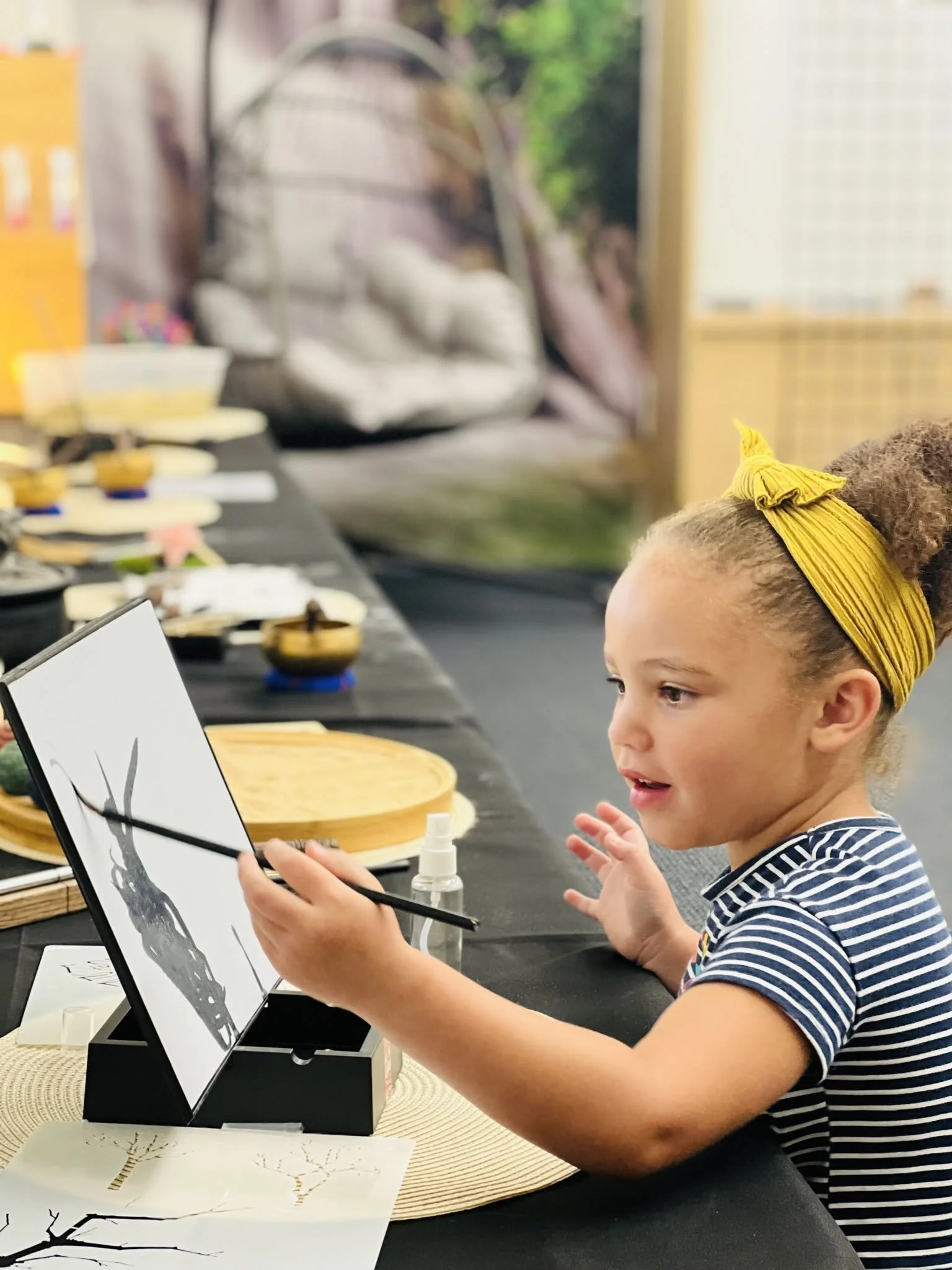 A young girl with curly hair, wearing a yellow headband and a striped shirt, is sitting at a table, holding a paintbrush and painting a black and white picture of a tree on a piece of paper. There are various art supplies and bowls on the table.