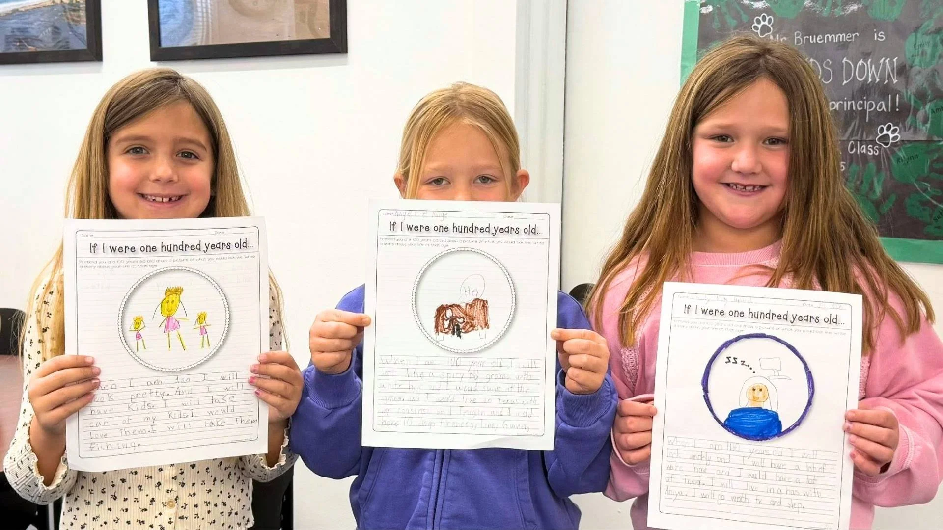 Three young girls are standing in a classroom holding up their writing projects. Each girl has a drawing on her paper, with the girl on the left having a picture of three stick figure girls, the girl in the middle drawing a brown animal with a speech bubble, and the girl on the right featuring a drawing of a yellow face inside a blue circle.