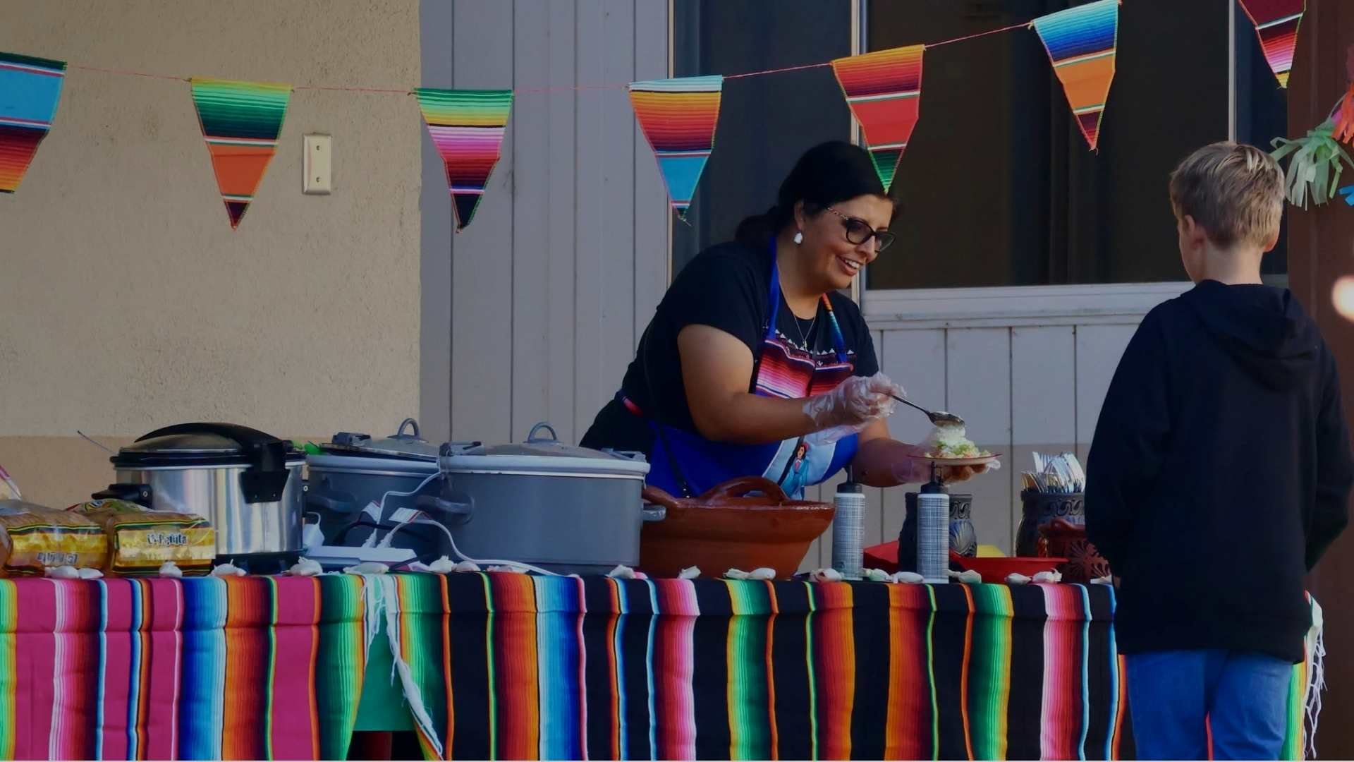 A woman preparing food at a festive outdoor event, with colorful striped decorations on the table and hanging banners in the background, and a young boy standing nearby.