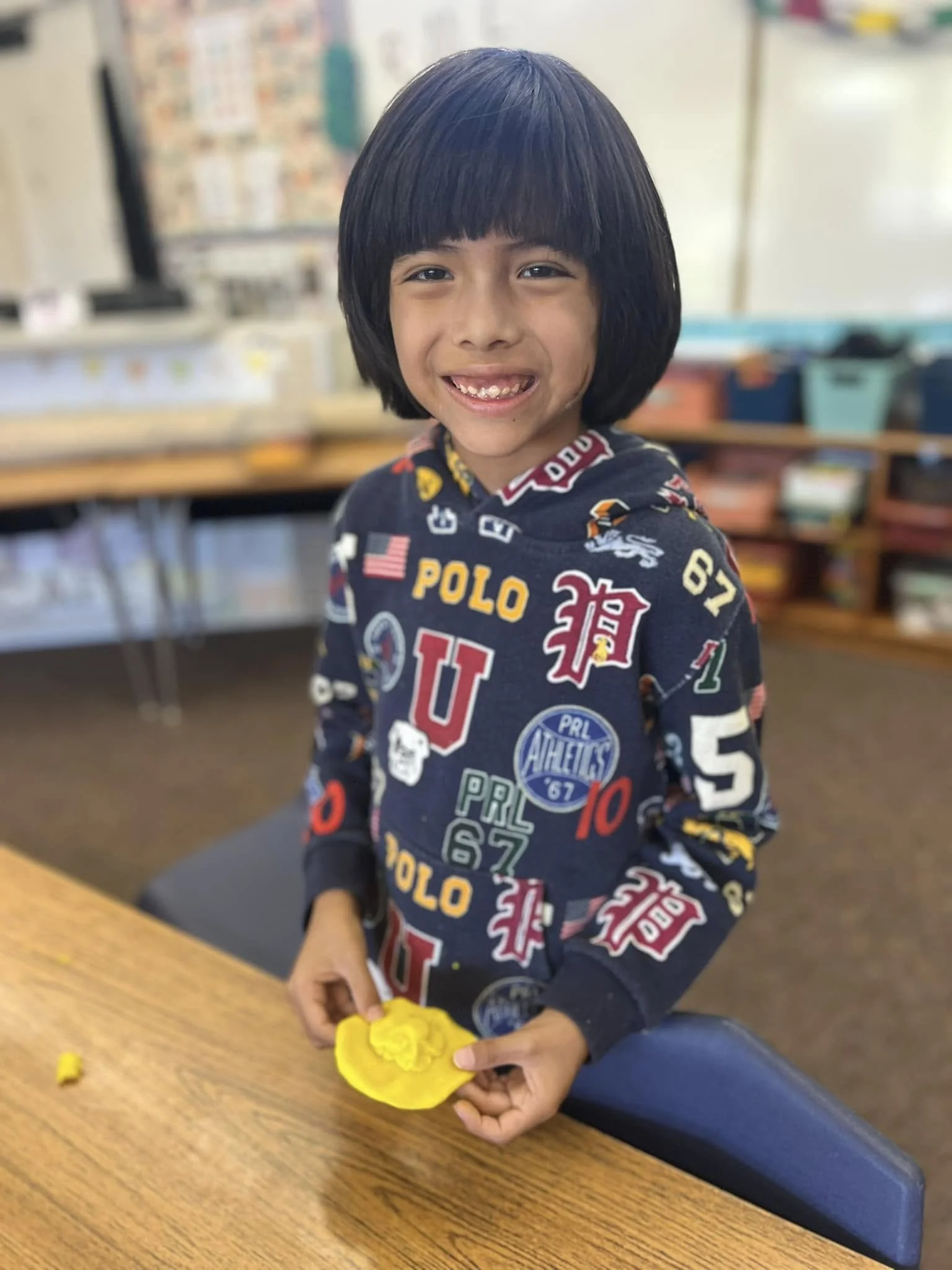 A young boy with dark hair and a big smile, wearing a navy sweatshirt with colorful patches and text, sitting at a wooden table in a classroom, holding a yellow slime. The background shows classroom supplies and a bulletin board.