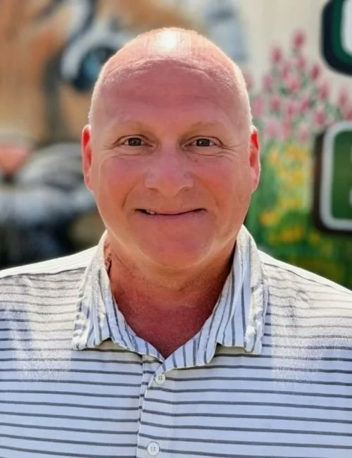 Superintendent Ian Trejo wearing a light-colored striped collared shirt, standing outdoors with a blurred colorful background.