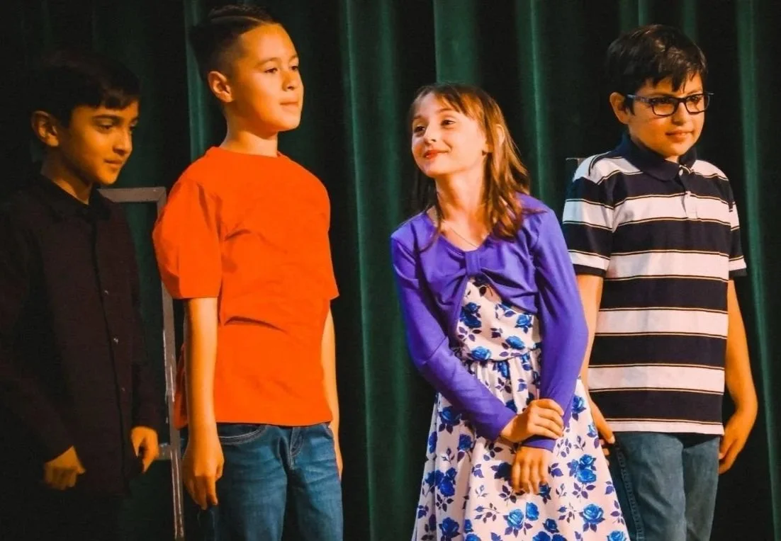 Four children standing in a line on stage with dark green curtains behind them. They are dressed casually and appear to be participating in a school performance or event.