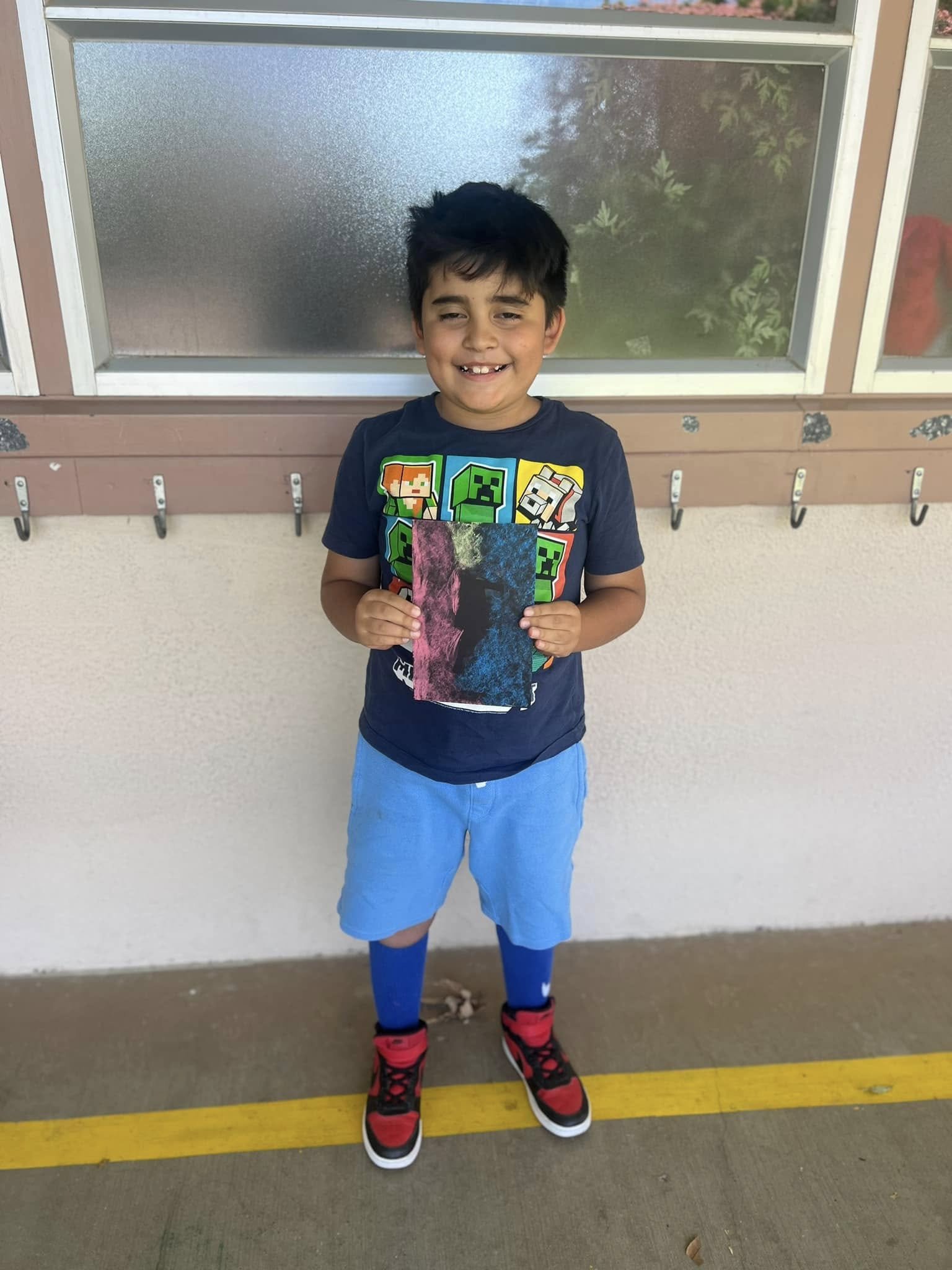A young boy with dark hair smiling and standing in front of a frosted glass window, holding a colorful piece of artwork, wearing a dark T-shirt with Minecraft characters, blue shorts, blue socks, and red and black sneakers.