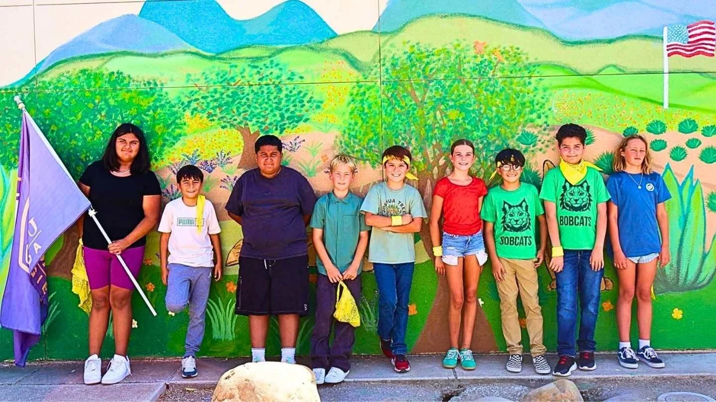 Group of children and a teenager standing in front of a colorful mural mural depicting a landscape with trees, hills, and mountains. Two children are wearing shirts with the Bobcat mascot and text.