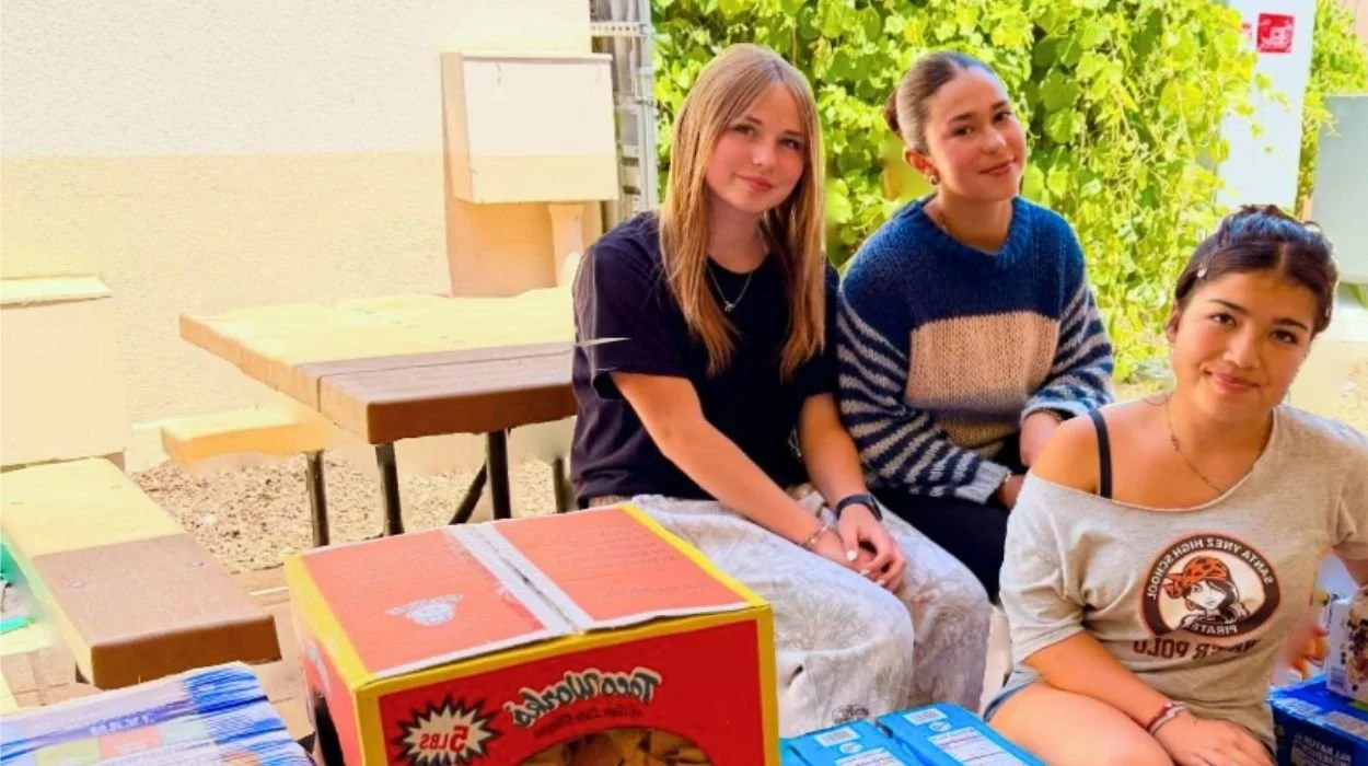 Three young women in middle school sitting at a table outdoors surrounded by food and snacks.