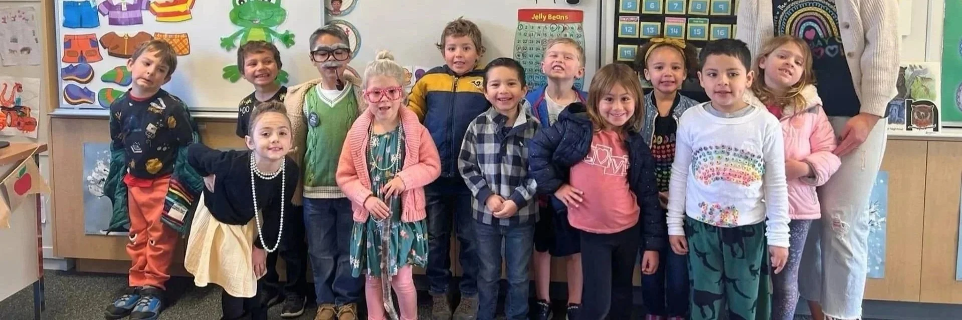 A group of young children standing together in a classroom, dressed in casual clothes, some wearing glasses and accessories, with educational posters and a whiteboard in the background.