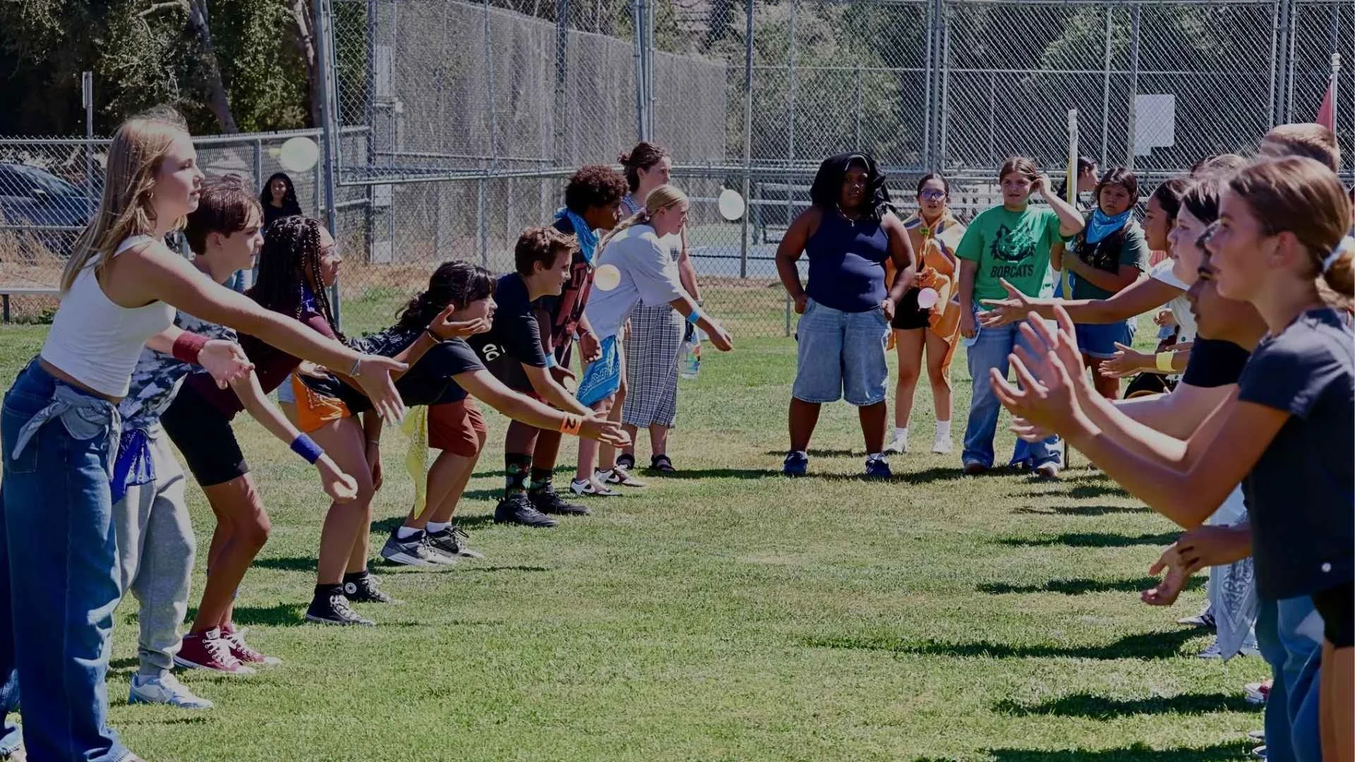 A group of children and adults on a sports field with a tall chain-link fence in the background. The children are lined up facing each other, engaging in an activity, while some adults stand nearby observing.