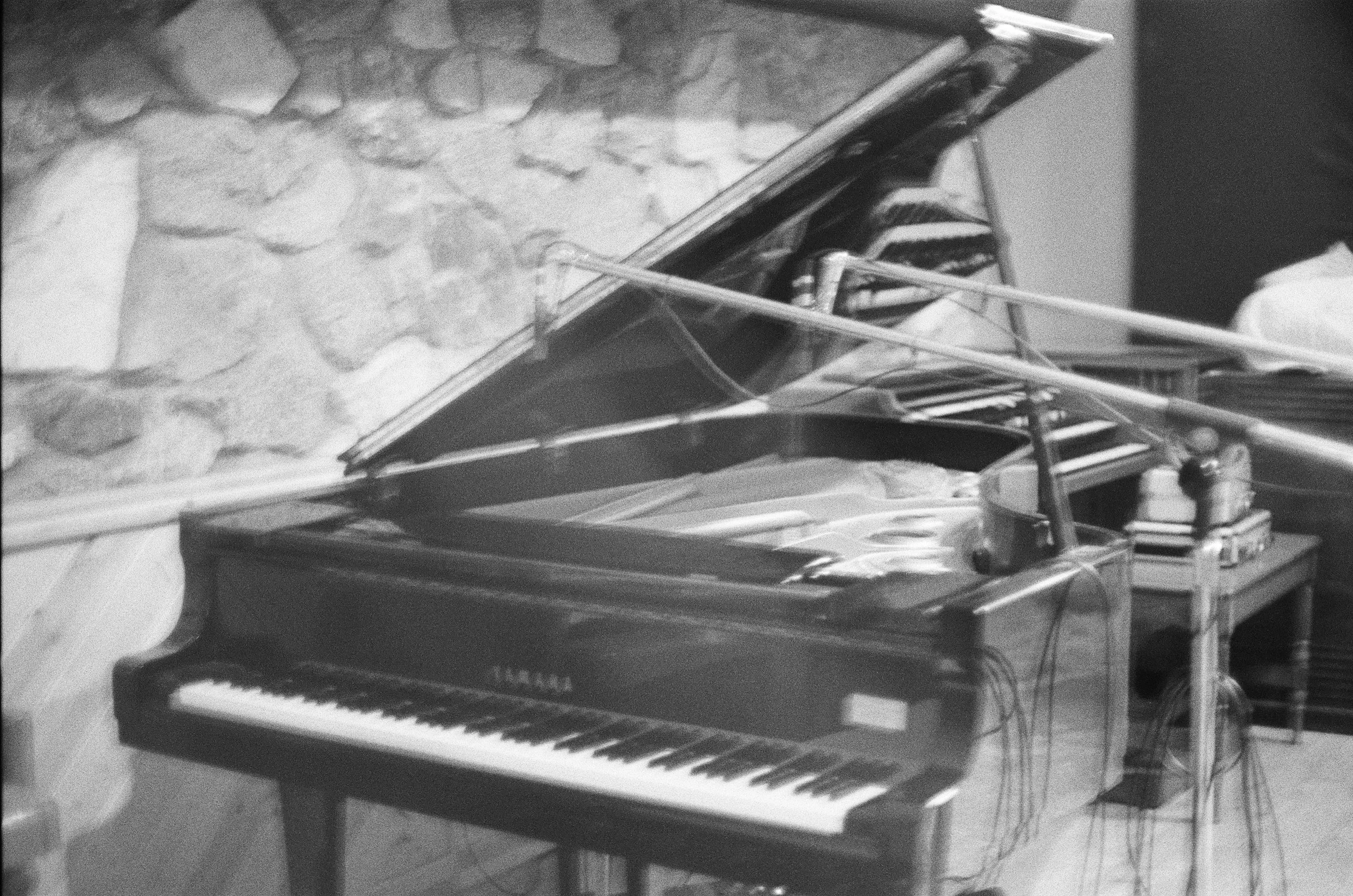 Black and white photo of a piano on a stand with a stack of books or sheet music on top, and an electric guitar leaning against the side of the stand, with a wall and some furniture visible in the background.