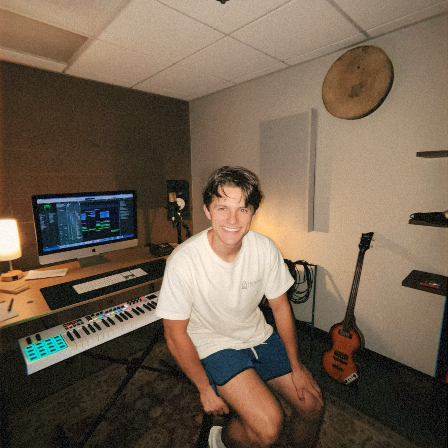 A young man with dark hair smiling, sitting in a music studio. He's wearing a white t-shirt and blue shorts. In front of him, there's a MIDI keyboard. To his left, a computer monitor displays a digital audio workstation. A guitar is placed against the wall to his right. The room has a brown and gray wall with a circular decoration above.